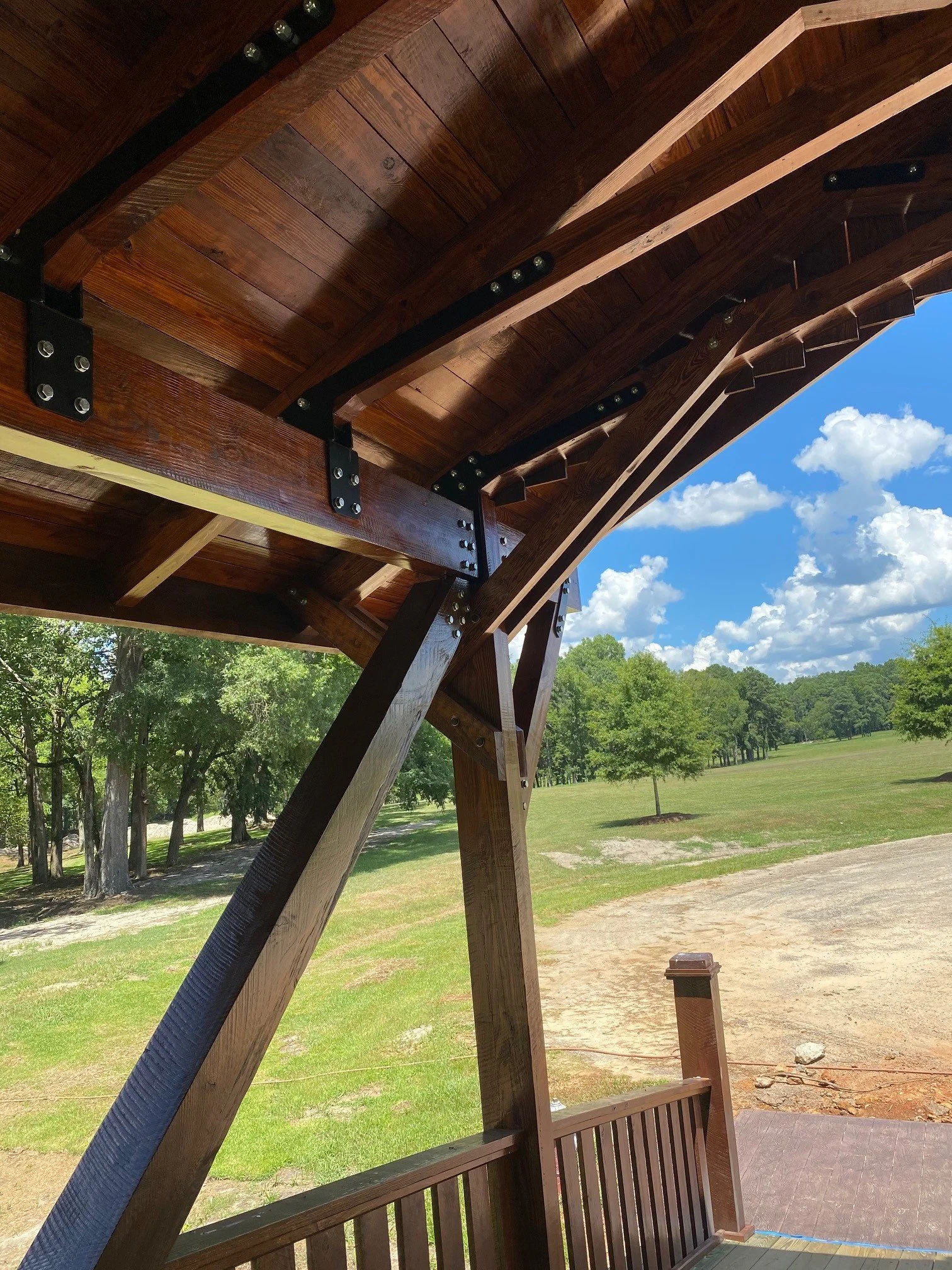 The underside of a wooden awning supported by metal brackets with bolts, shading a outdoor wooden deck area. Green grass and trees are visible in the background under a blue sky with scattered clouds.