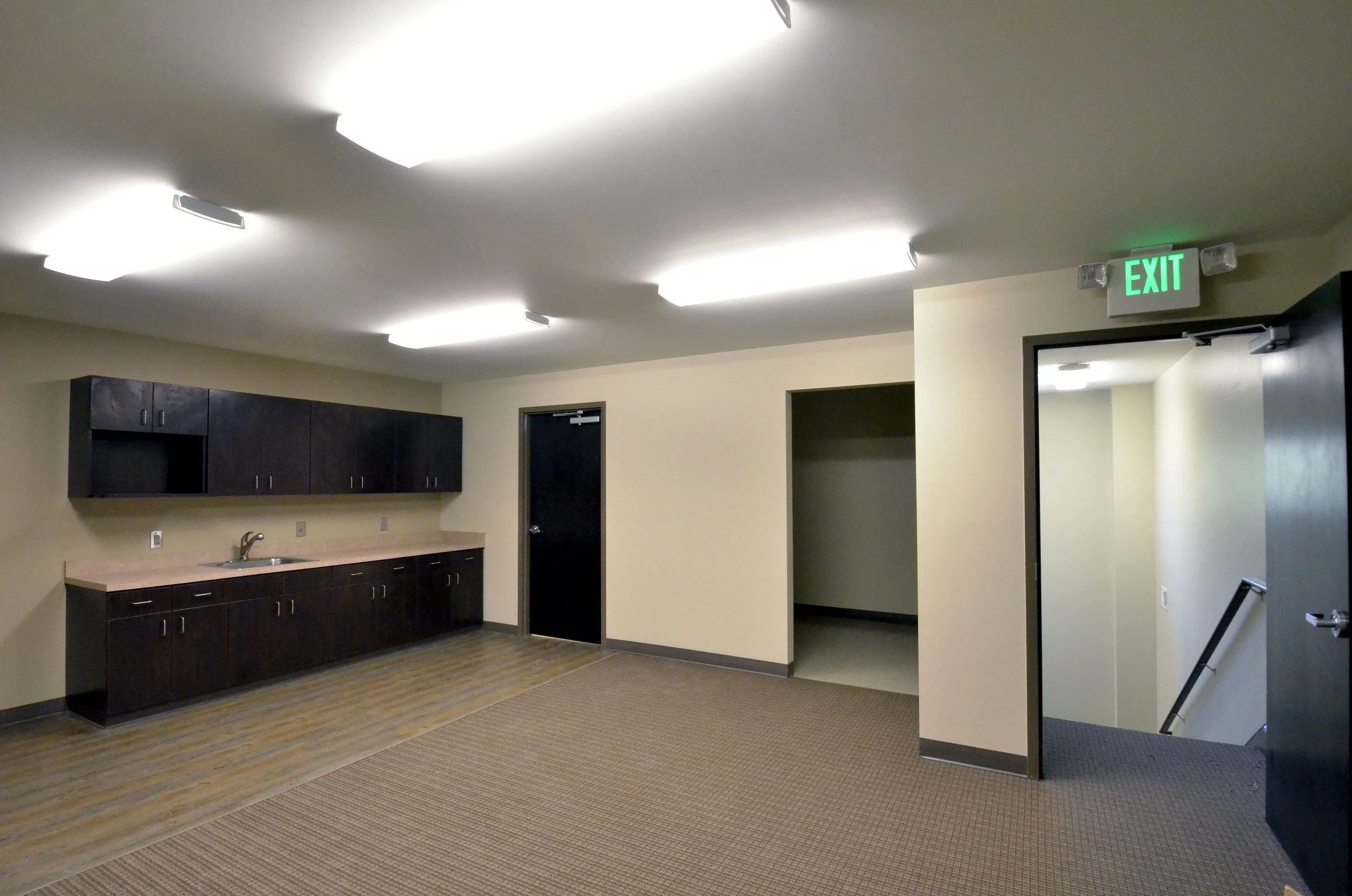 An indoor room with beige walls and ceiling, fluorescent lights, a door with an illuminated exit sign above it, dark-colored cabinets and countertop, and a carpeted floor.