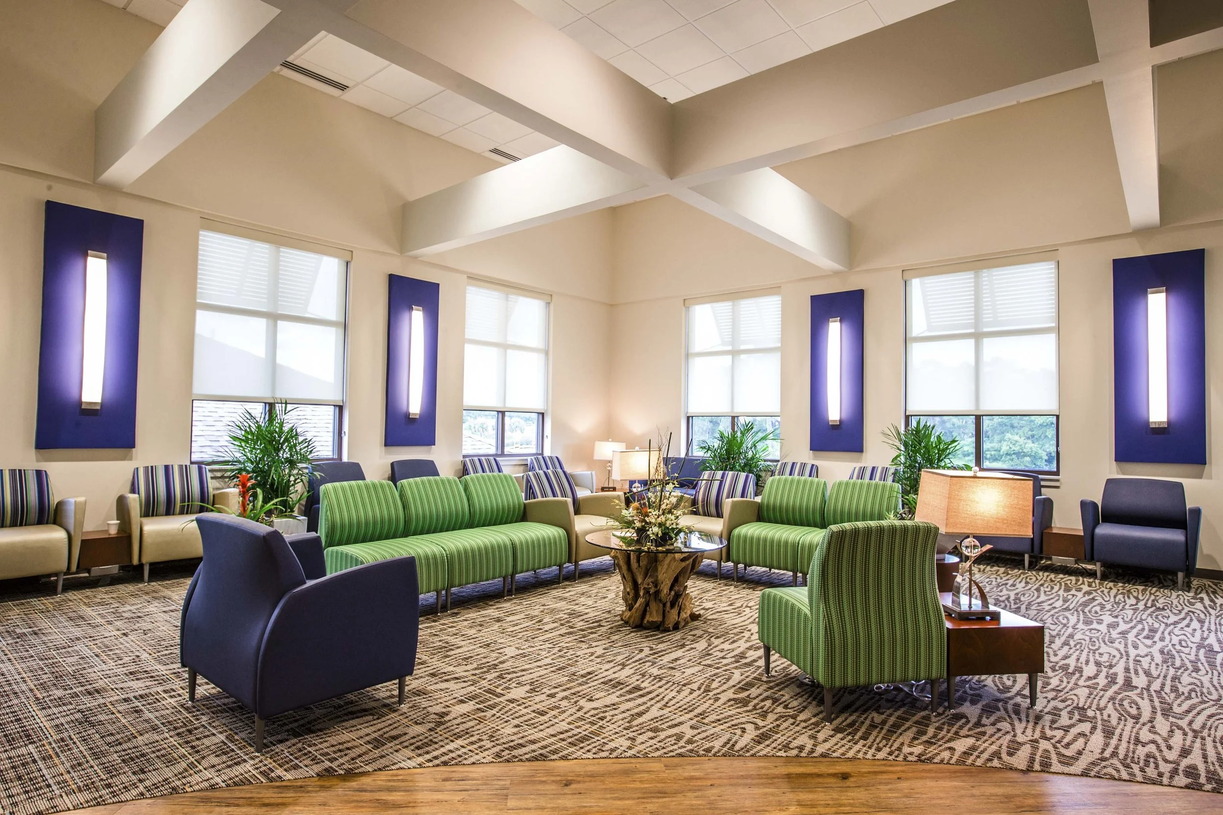 A spacious waiting room with green, purple, and beige chairs, large windows with blinds, potted plants, and decorative lighting on purple walls.