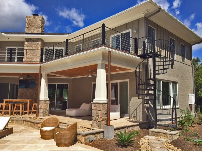 Exterior view of a modern two-story house with a balcony, outdoor seating area, and a spiral staircase on the side. The house features brick and siding exterior with black accents, a patio, and some gardening.