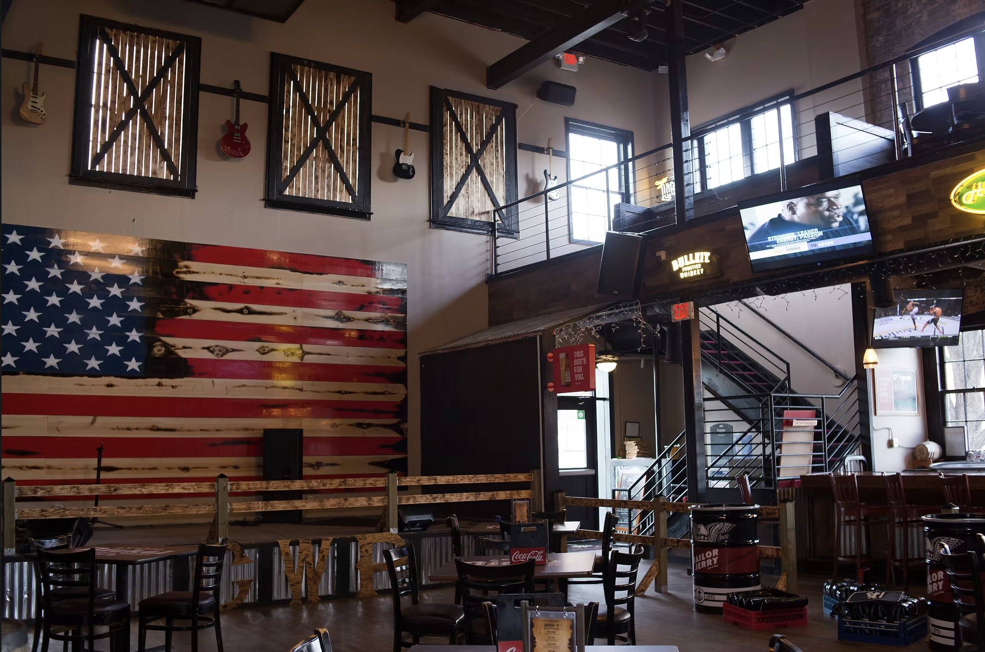 Interior of a restaurant or bar with wooden tables and chairs, a large American flag wall art, guitars on the wall, multiple television screens showing sports, and a staircase leading to an upper level with windows and decor.
