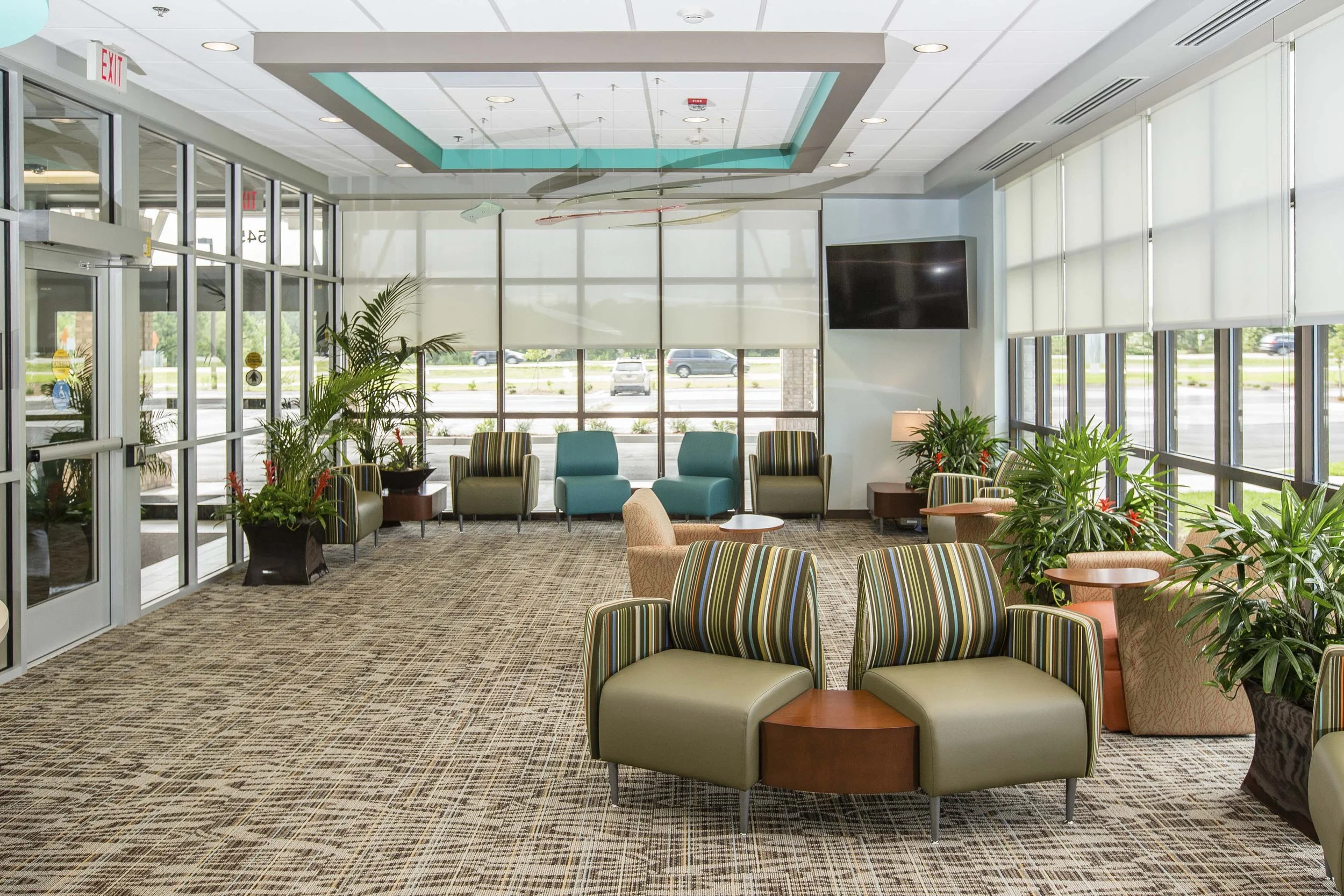 A modern hospital or clinic waiting area with large windows, several potted plants, colorful striped chairs, a television mounted on the wall, and a glass door entrance.
