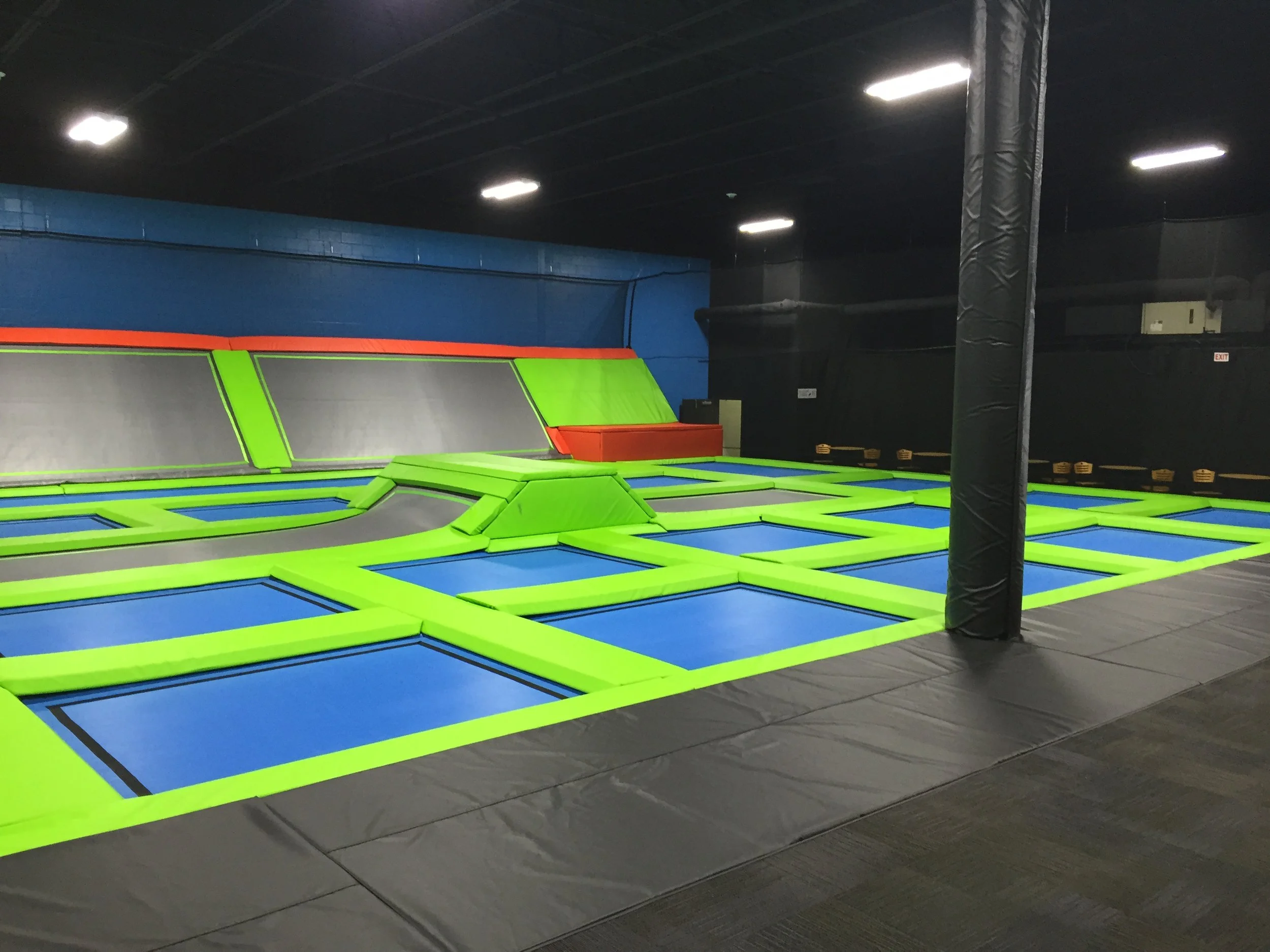 Indoor trampoline park with colorful padded trampolines, black wall, blue wall, and ceiling with bright overhead lights.