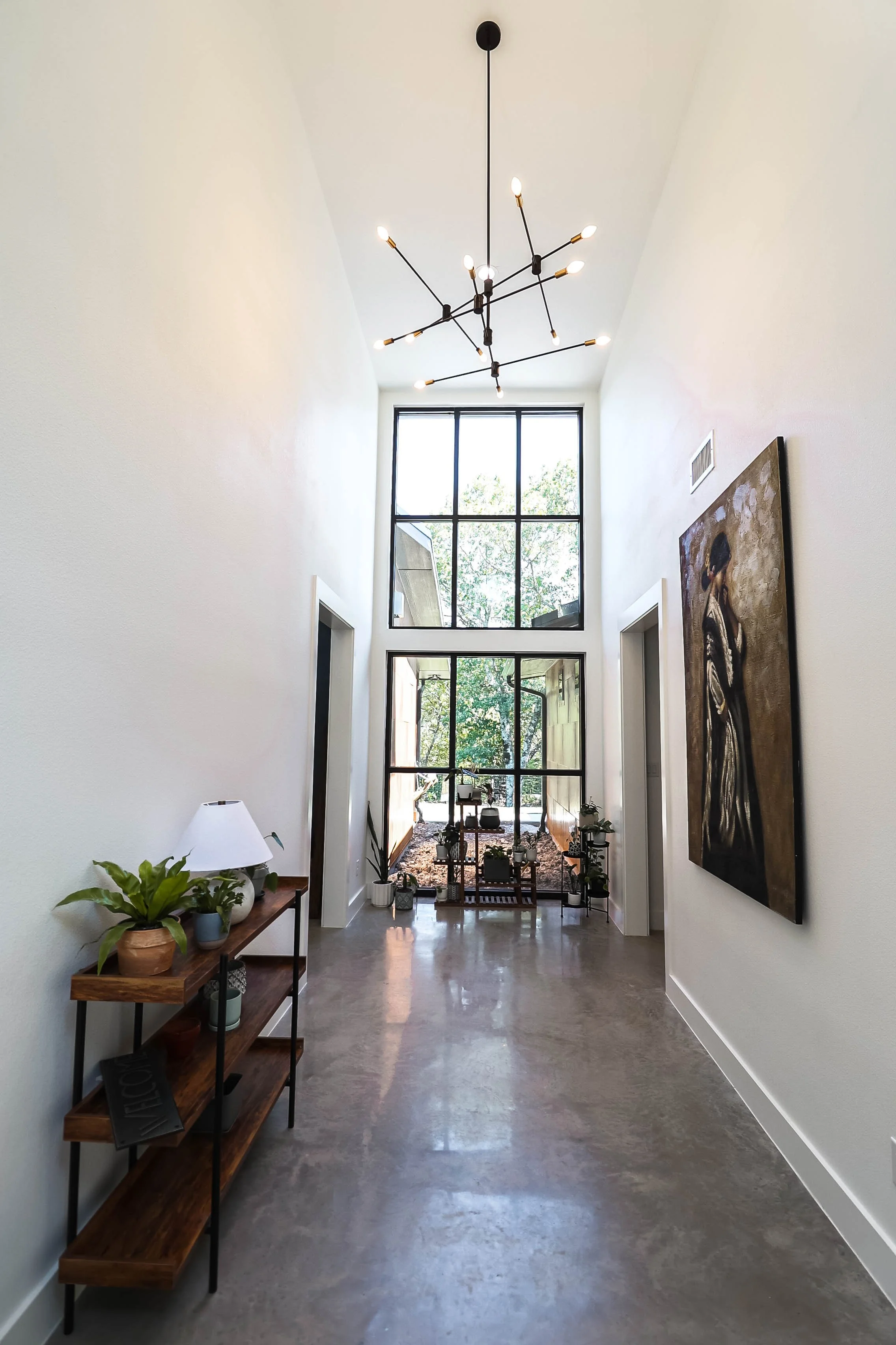 A bright, modern hallway with large floor-to-ceiling windows at the end, a black geometric chandelier hanging from the ceiling, and a wooden and metal console table on the left decorated with potted plants and a table lamp. A large abstract painting 
