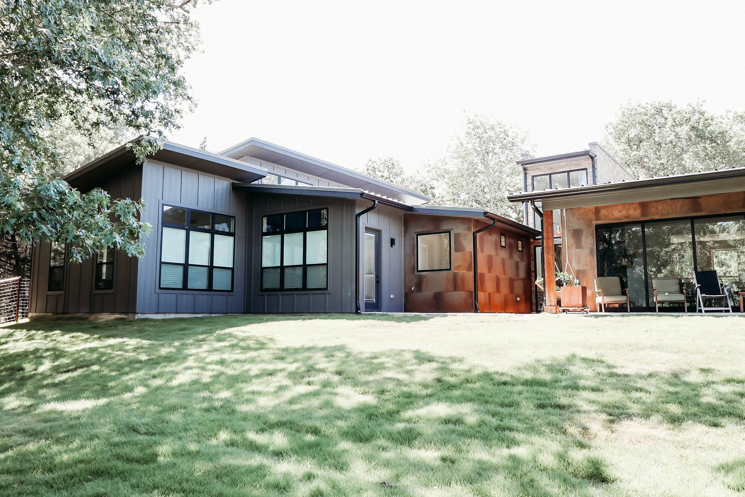 Modern house with large windows, metal and stone exterior, and a backyard with green grass and patio furniture.