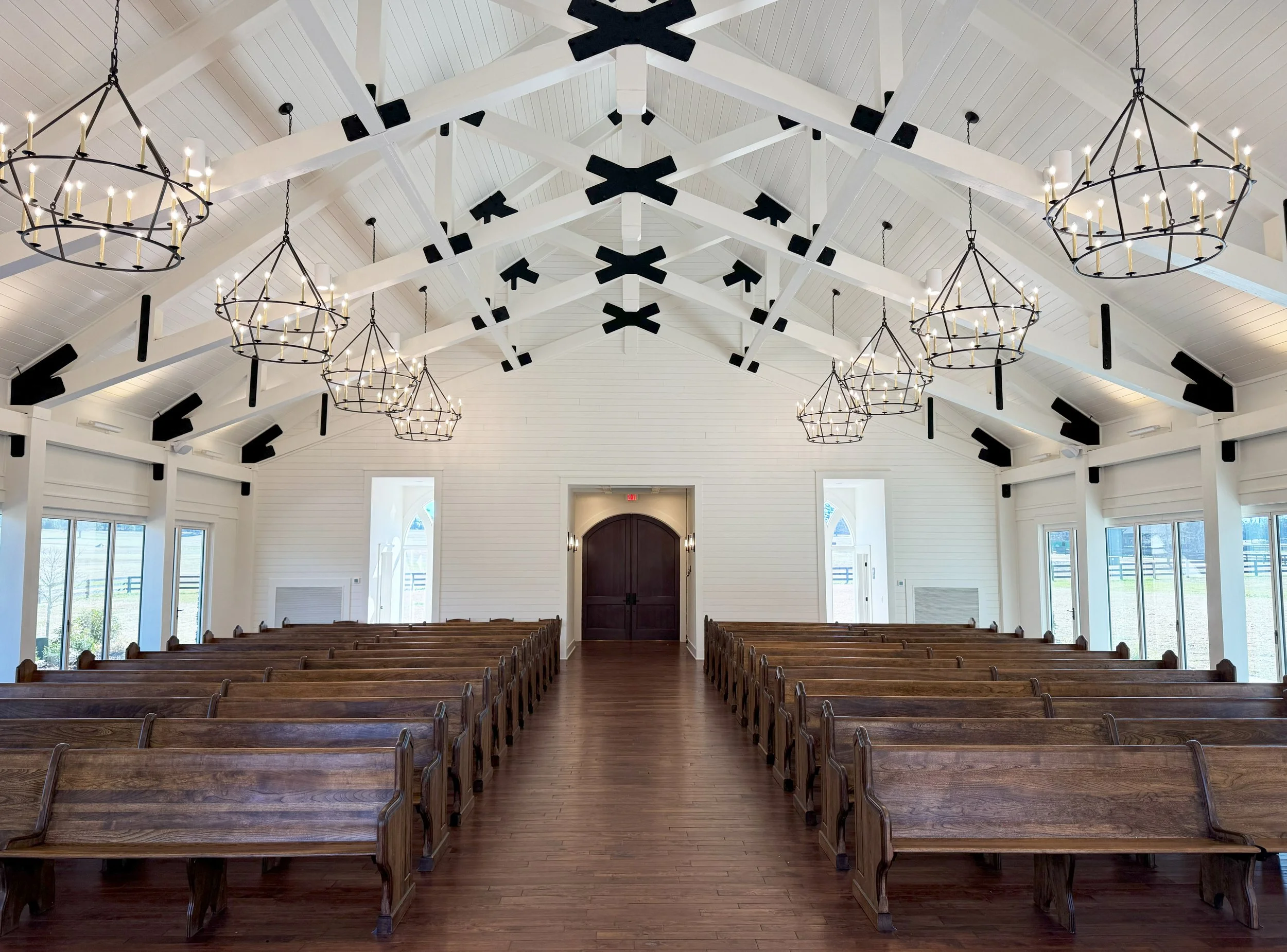 Interior of a church or chapel with wooden pews, white walls, high vaulted ceiling with exposed beams, black accents, and multiple chandeliers with candles hanging from the ceiling.