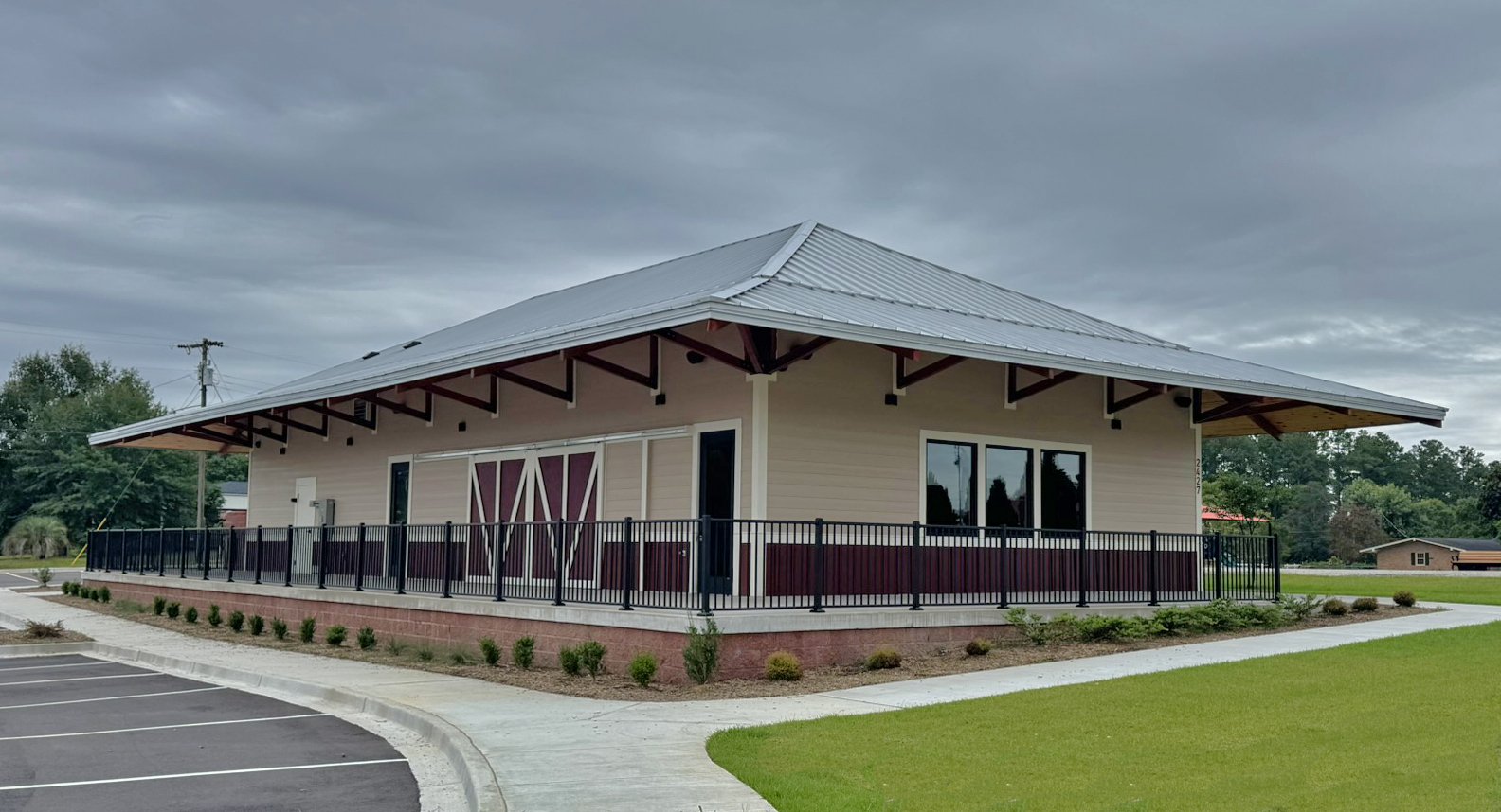 A modern building with a metal roof, surrounded by a black metal railing, located in a grassy area with parking spaces nearby under a cloudy sky.