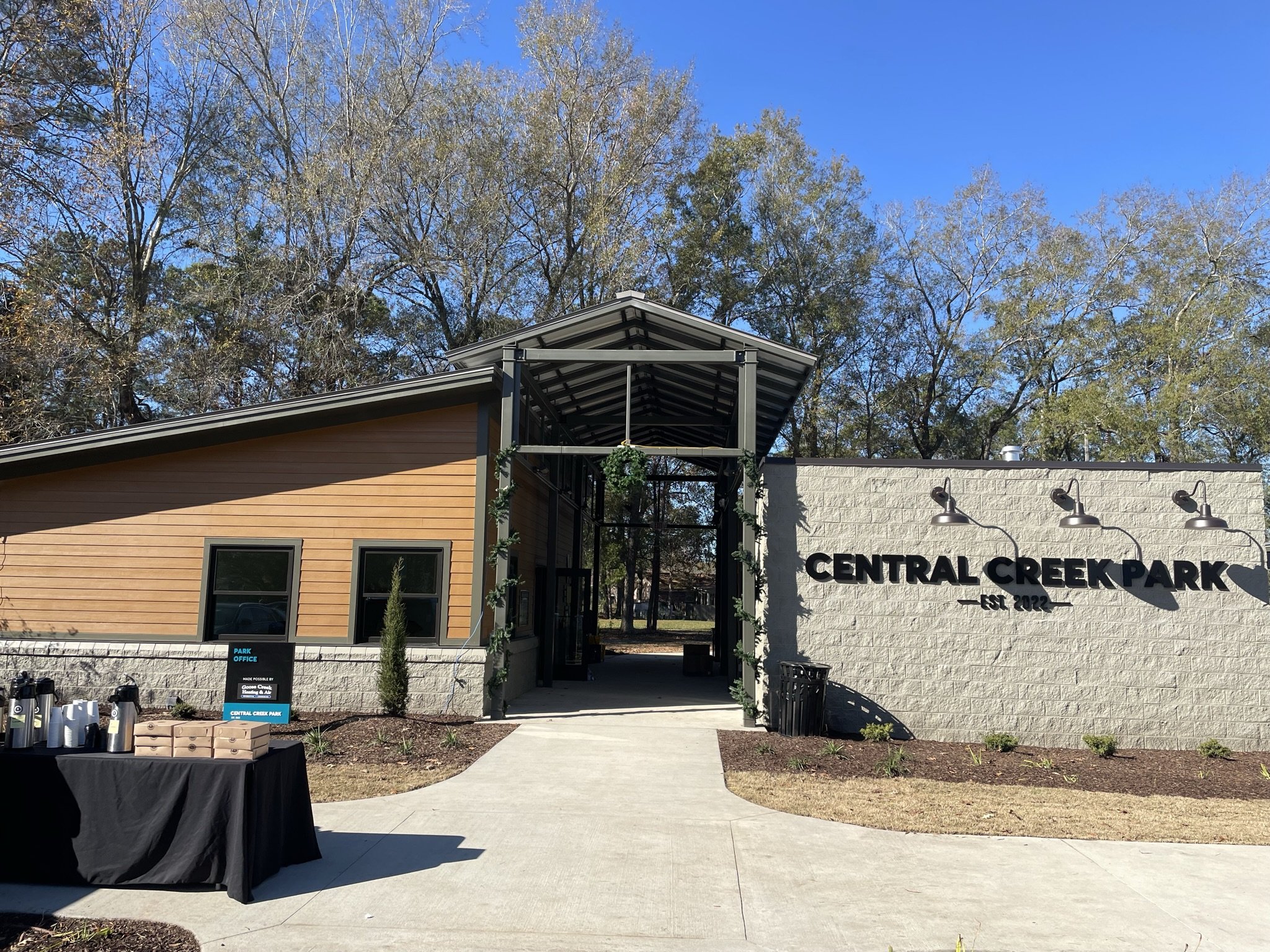 Entrance to Central Creek Park with a sign that says 'EST 2022'. Modern building with wooden siding, metal roof, and outdoor lighting fixtures. Clear sky and trees in the background.