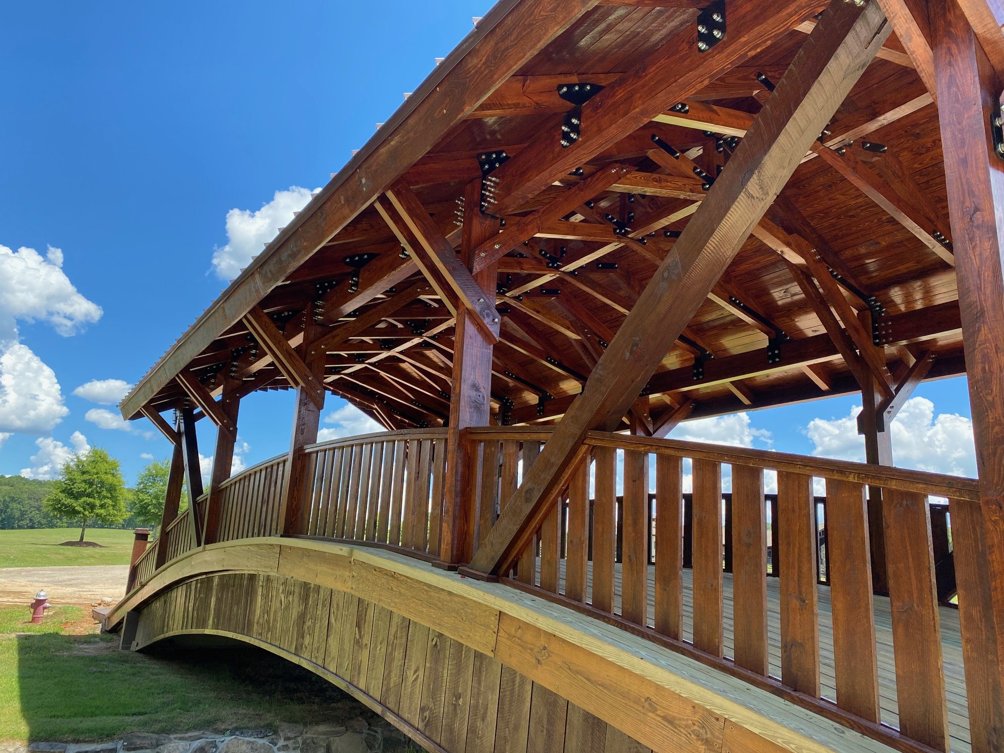Wooden bridge structure with curved design, under a partly cloudy blue sky, set in a park with grassy area and trees in the background.