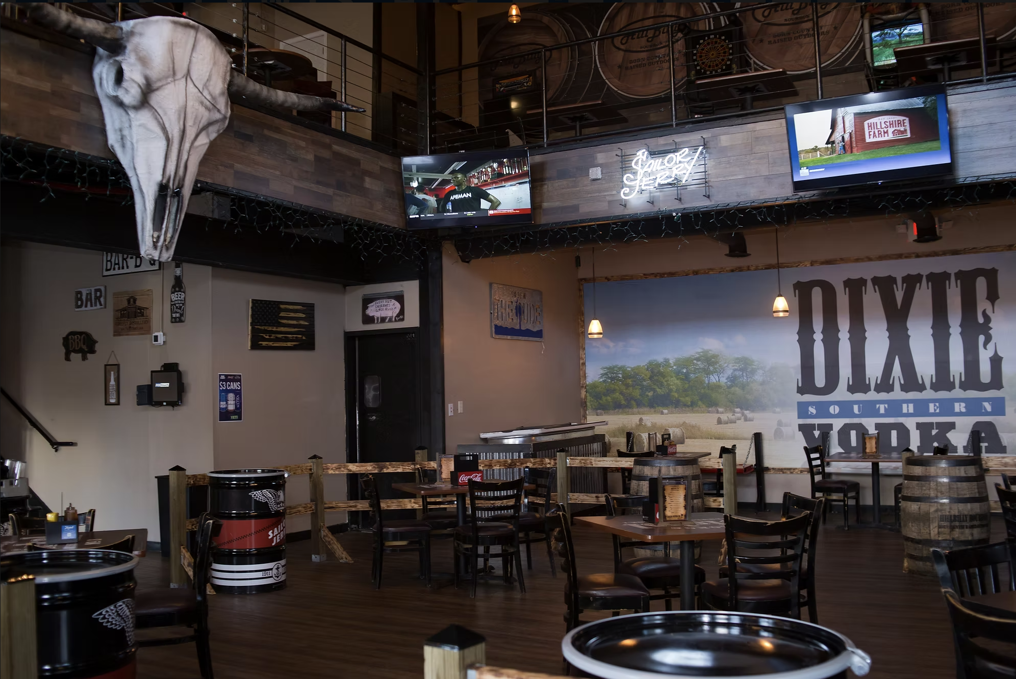 Interior of a Southern-style restaurant with wooden decor, multiple tables and chairs, a large mural of rural farmland, a mounted longhorn skull, several wall signs, and television screens showing different programs.