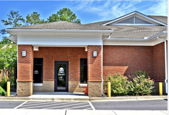 Front of a brick commercial building with glass door and windows, surrounded by small bushes, yellow posts, and a parking lot, under a blue sky with some clouds.