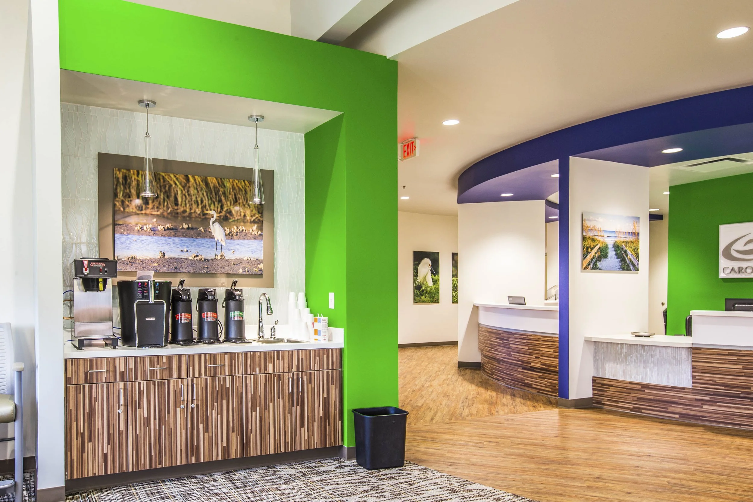 Interior of a modern cafe or reception area with vibrant green, blue, and white walls, wooden accents, ceiling lights, a coffee station, and framed nature photographs.