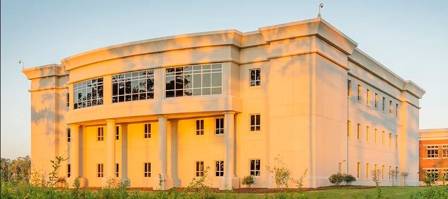 A large, modern beige building with tall columns and multiple windows, illuminated by the warm glow of sunset, surrounded by small trees and grass.