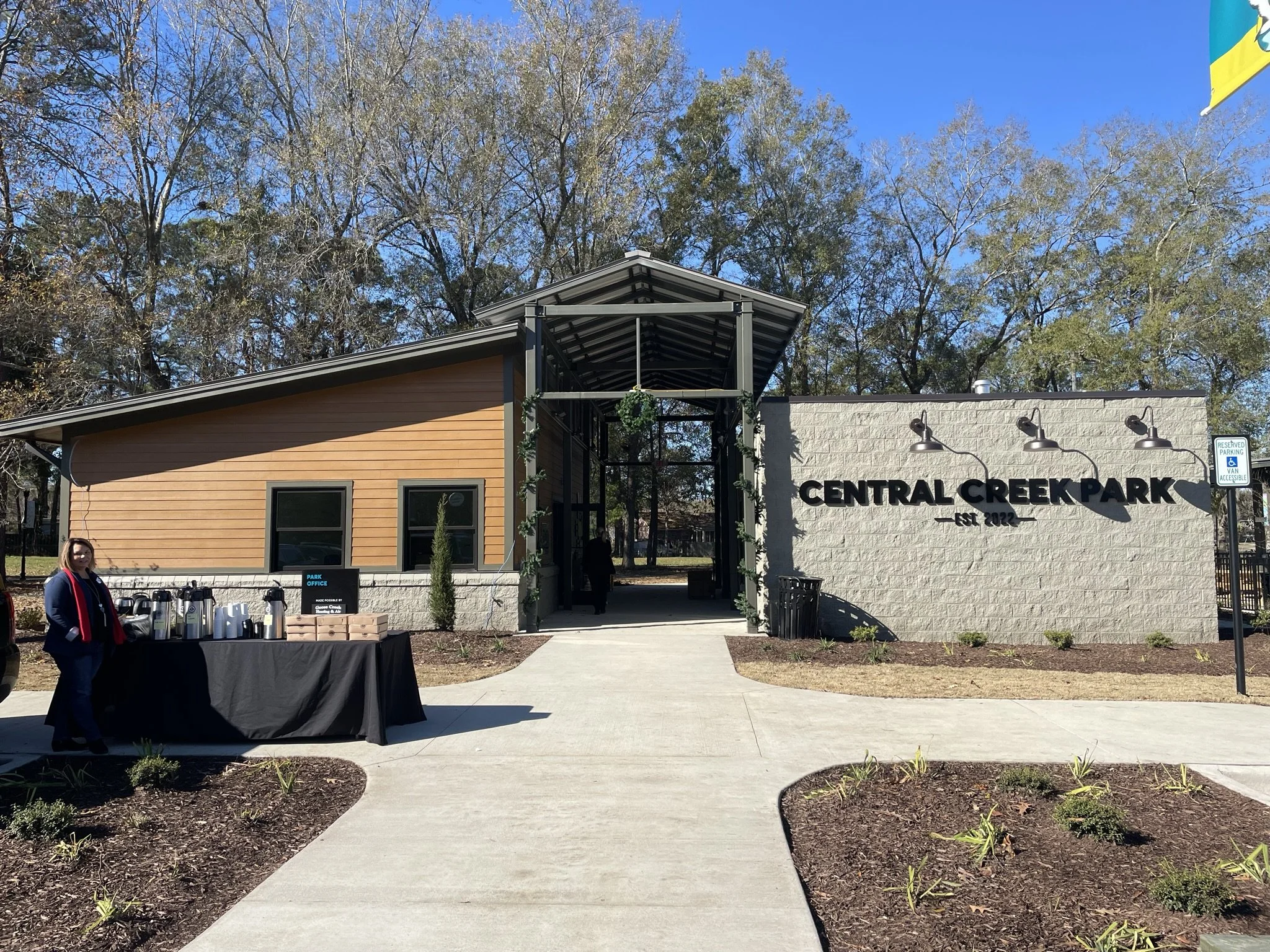 Entrance to Central Creek Park with a sign, a building with a partly wooden exterior, and an outdoor table with beverages on a black cover. There are trees and a blue sky in the background.
