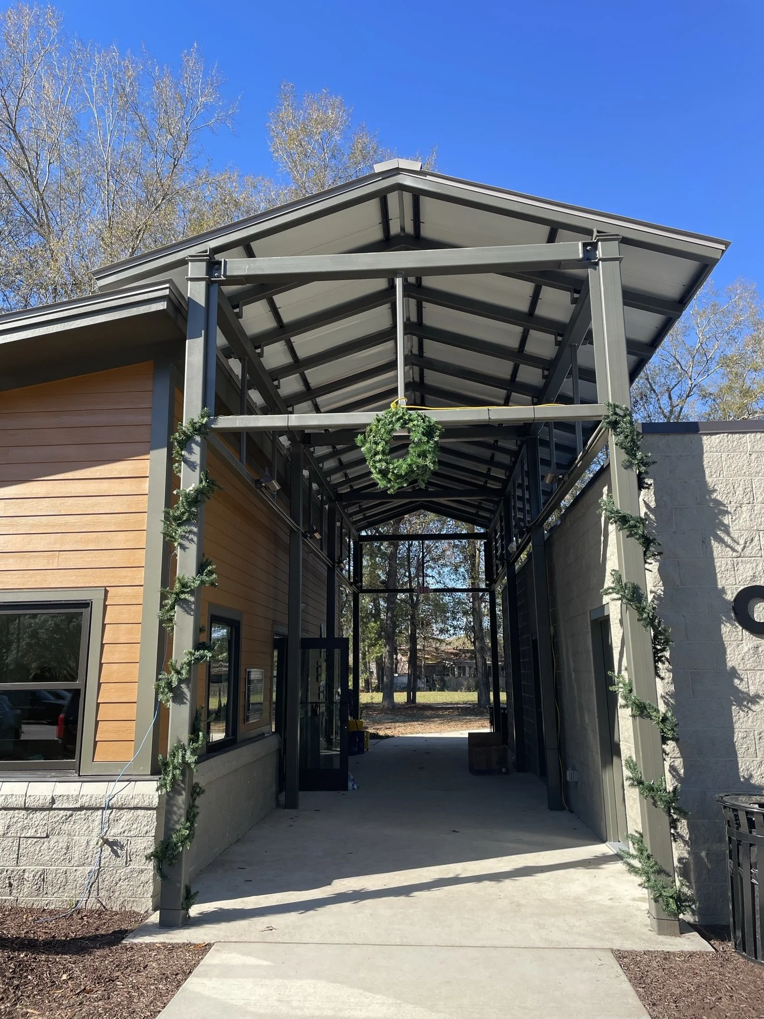 Newly constructed building with a metal and wood exterior, decorated with holiday greenery and a wreath, under a clear blue sky.