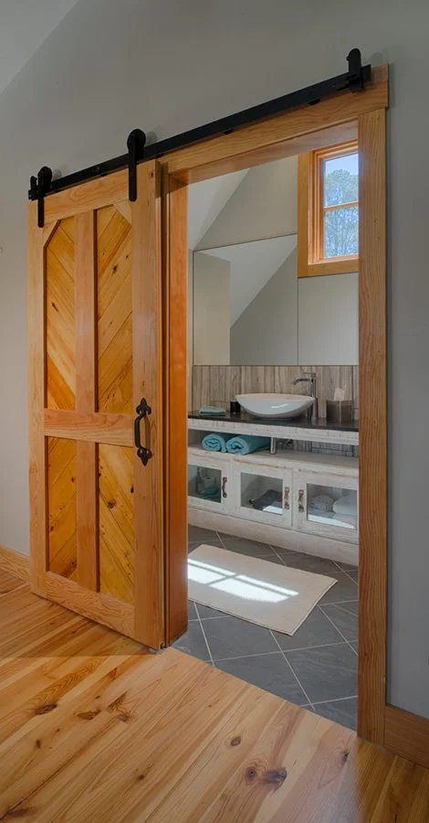 Wooden sliding barn door leading to a bathroom with a vessel sink, mirror, and towel shelf, illuminated by natural light from a small window.