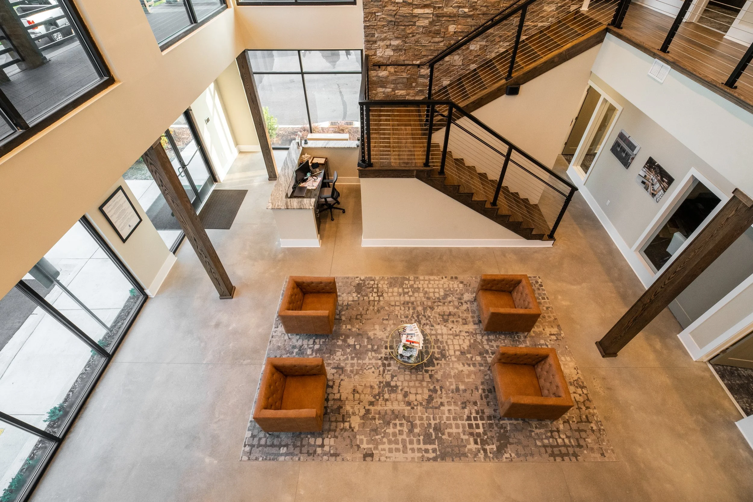 Interior view of a modern lobby with a staircase, glass walls, a reception desk, seating area with brown chairs, and artwork on the walls.