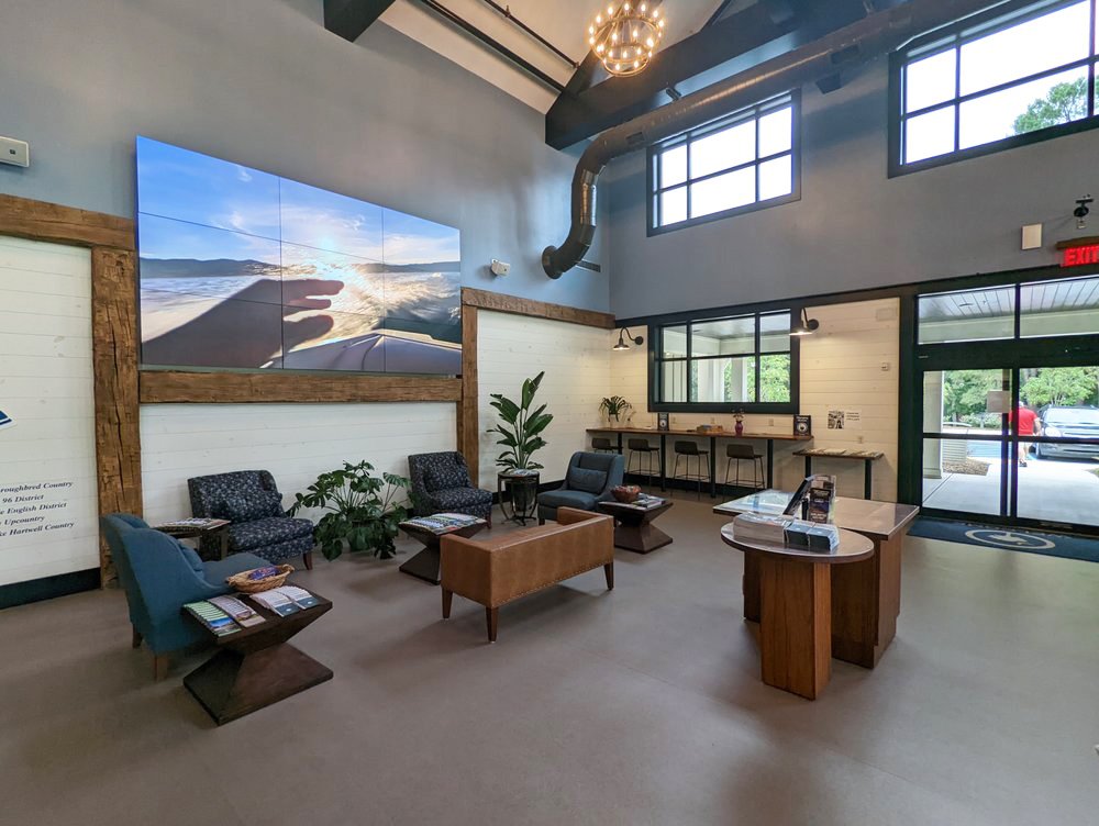 A modern lobby with high ceilings, large windows, and a wall-mounted digital screen displaying a scenic ocean view. The space includes seating with armchairs and a sofa, a wooden coffee table, potted plants, and a reception desk. There are high barst