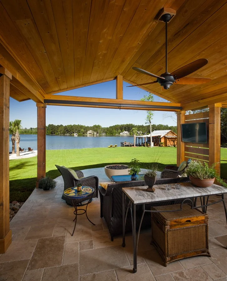 Covered outdoor patio with a fan, overlooking a lake with trees and boats, with patio furniture, potted plants, and a scenic view.