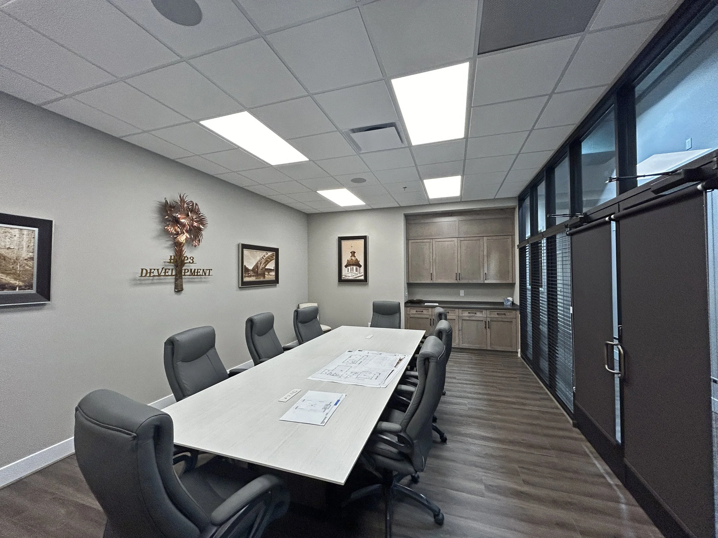 A conference room with a long table and multiple office chairs around it. The room has a gray wall with framed pictures and a decorative wall hanging. The ceiling has bright rectangular lights, and there are large windows with blinds on one side.
