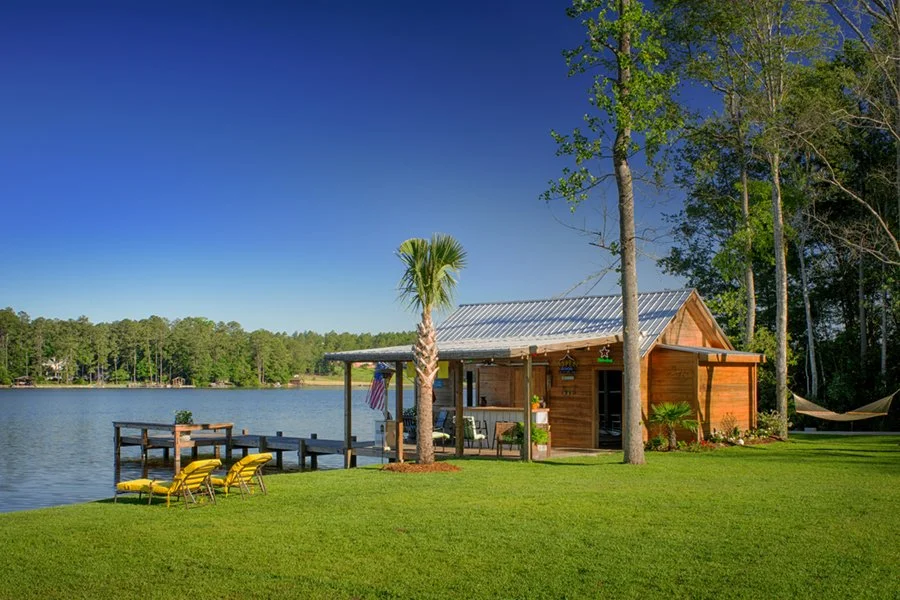 A lakeside scene with a wooden cabin, palm trees, green grass, yellow lounge chairs, and a dock extending into the lake, with a hammock on the right side and a bright blue sky.