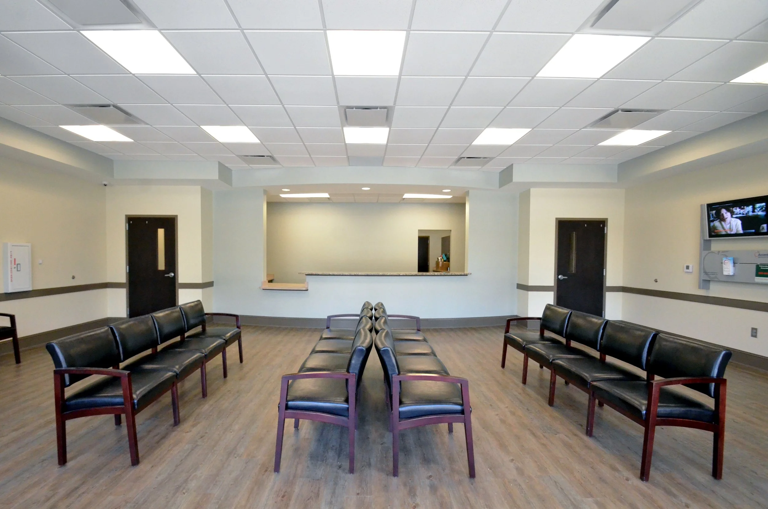 Empty waiting room with rows of black chairs, a reception window, a TV on the wall, and a wooden floor.