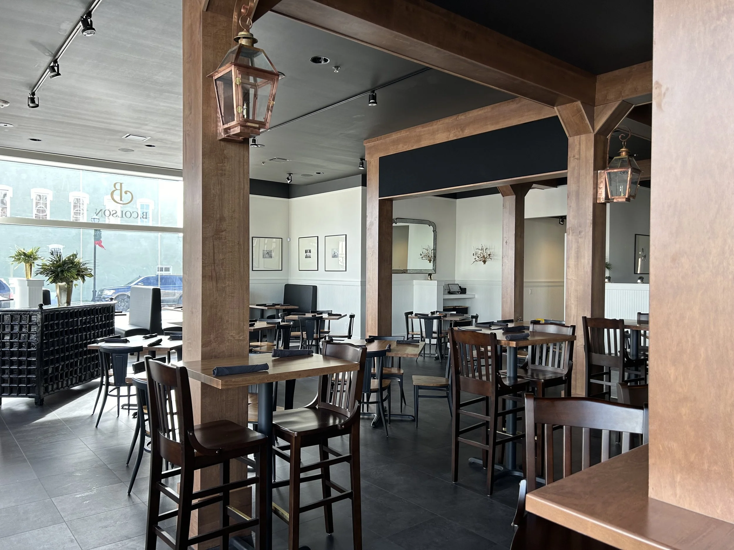 Interior of a modern restaurant with wooden beams, black tables and chairs, and hanging lantern-style lights, with large windows letting in natural light.