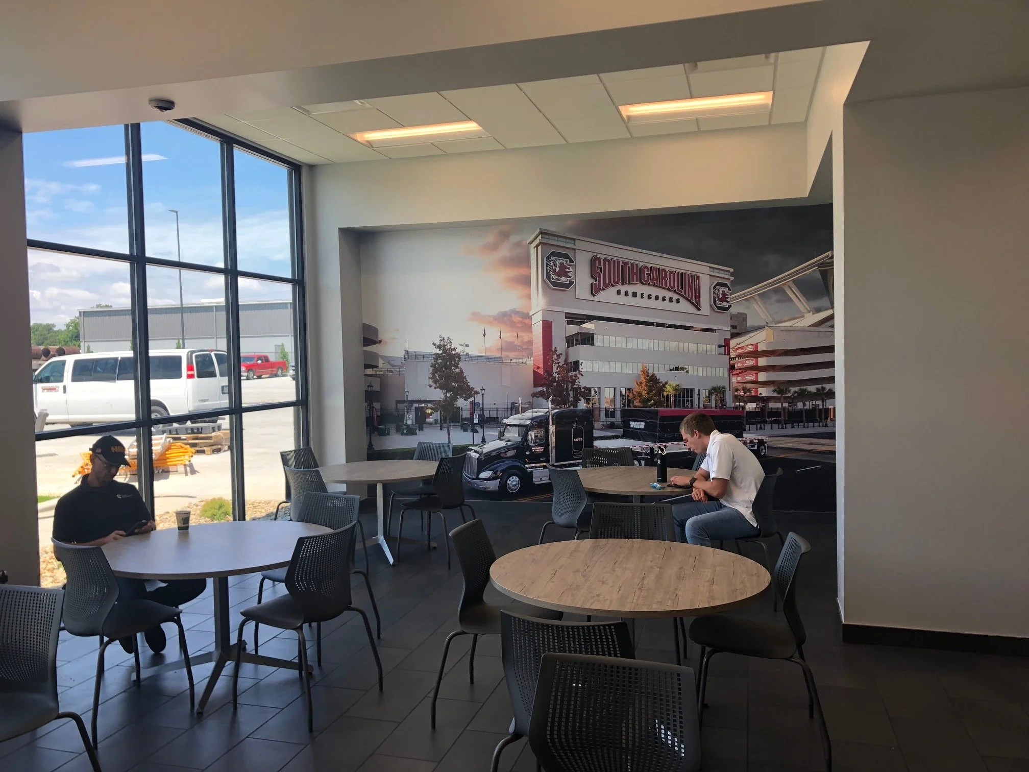 View of the interior of a building with round tables and chairs, showing large windows revealing an outdoor parking lot with vehicles and a South Carolina Gamecocks football stadium photo on the wall.