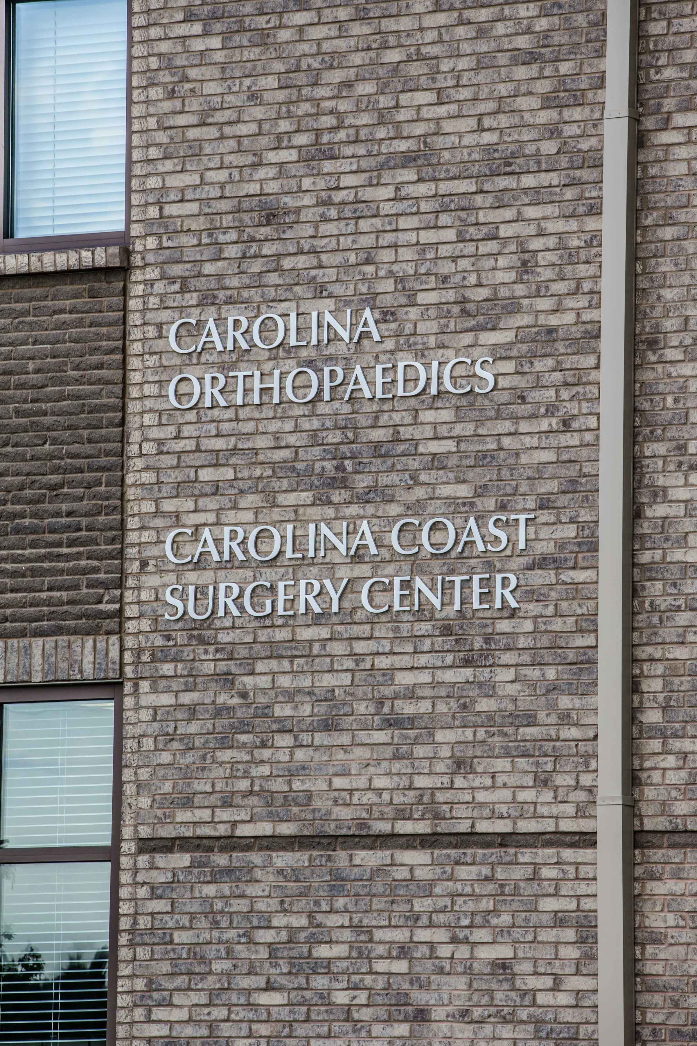Sign on a brick building reading 'Carolina Orthopaedics' and 'Carolina Coast Surgery Center' with partially visible windows.