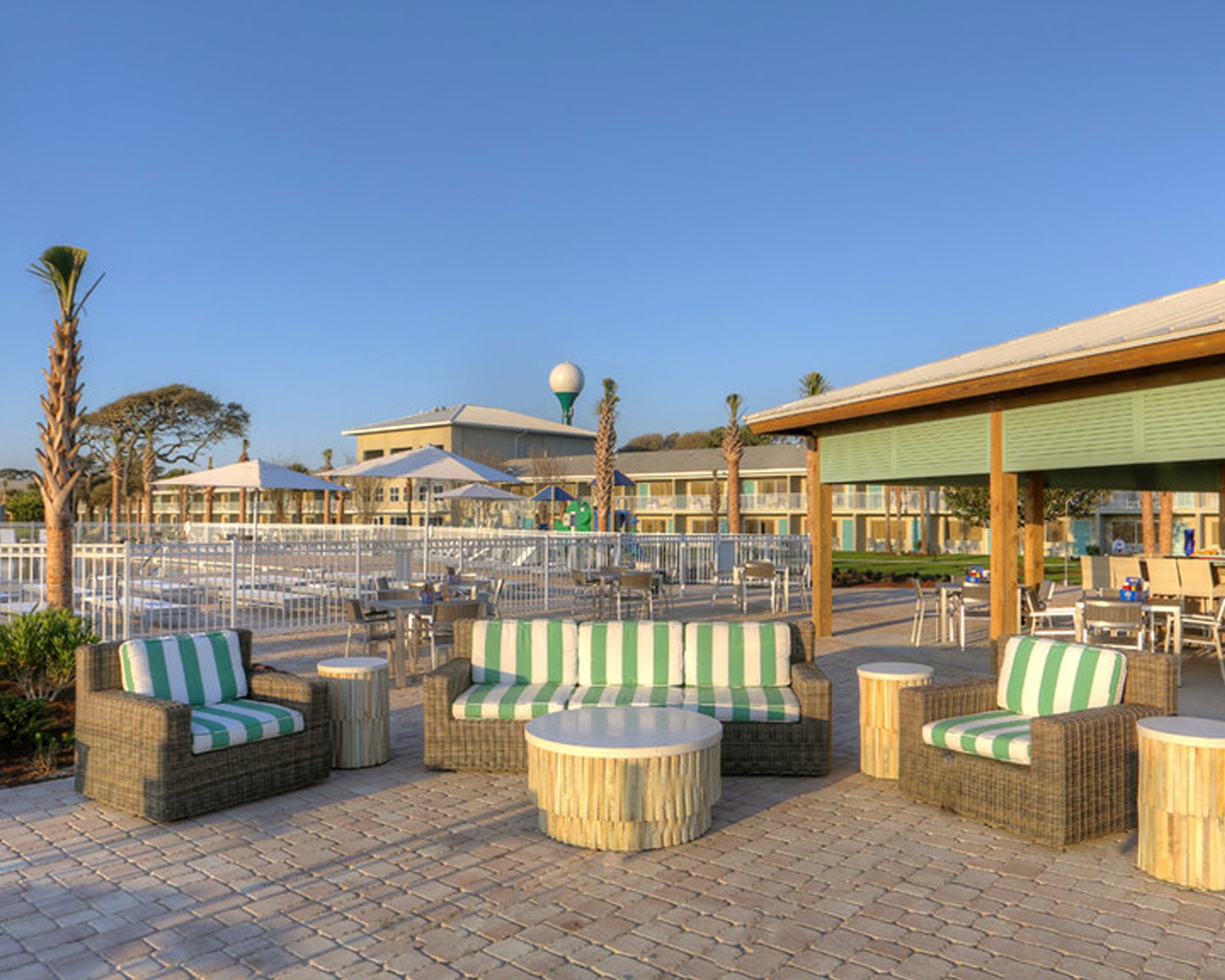 Outdoor seating area with wicker furniture with green and white striped cushions, round tables, wooden pillars, and a clear blue sky. In the background, a building with multiple floors, trees, and a water tower are visible.