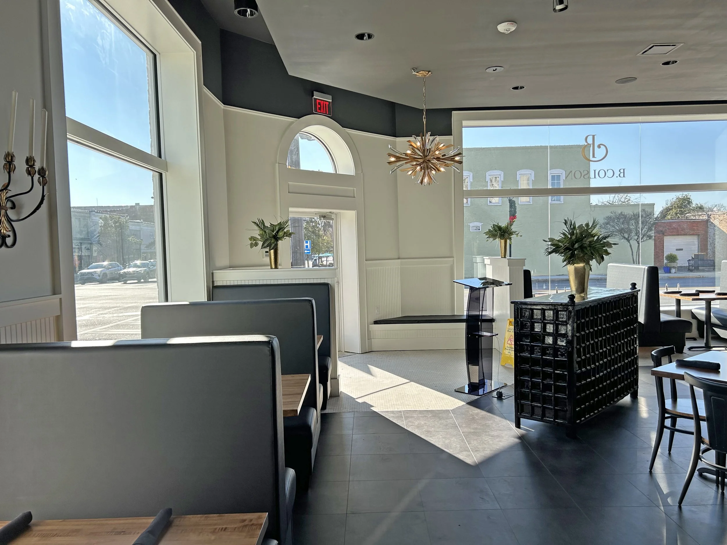 Inside a modern restaurant or café with large windows, stylish chandelier, potted plants on white ledges, and various tables and chairs, some with napkins and table settings. Bright daylight outside, a parking lot, and colorful building can be seen t