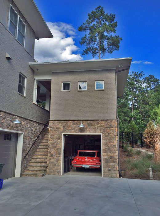A two-story house with a stone garage entrance, a red vintage car inside, stone stairs on the left, and small windows on the second floor, with trees and blue sky in the background.