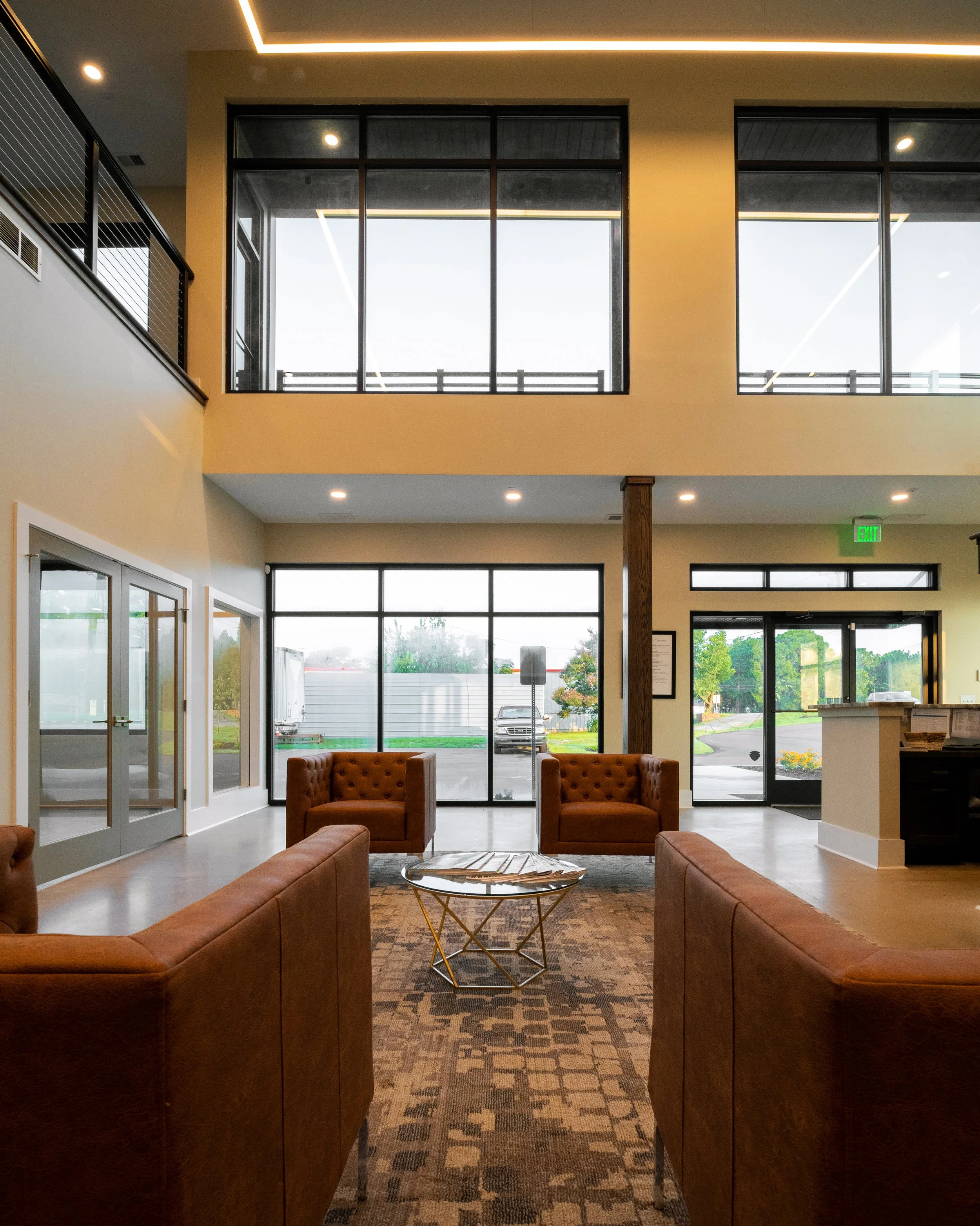 A spacious hotel lobby with brown leather couches arranged in a U-shape, a round glass coffee table, large floor-to-ceiling windows showing outside scenery, and a high ceiling with modern lighting. The interior features a patterned rug and neutral-colored walls.