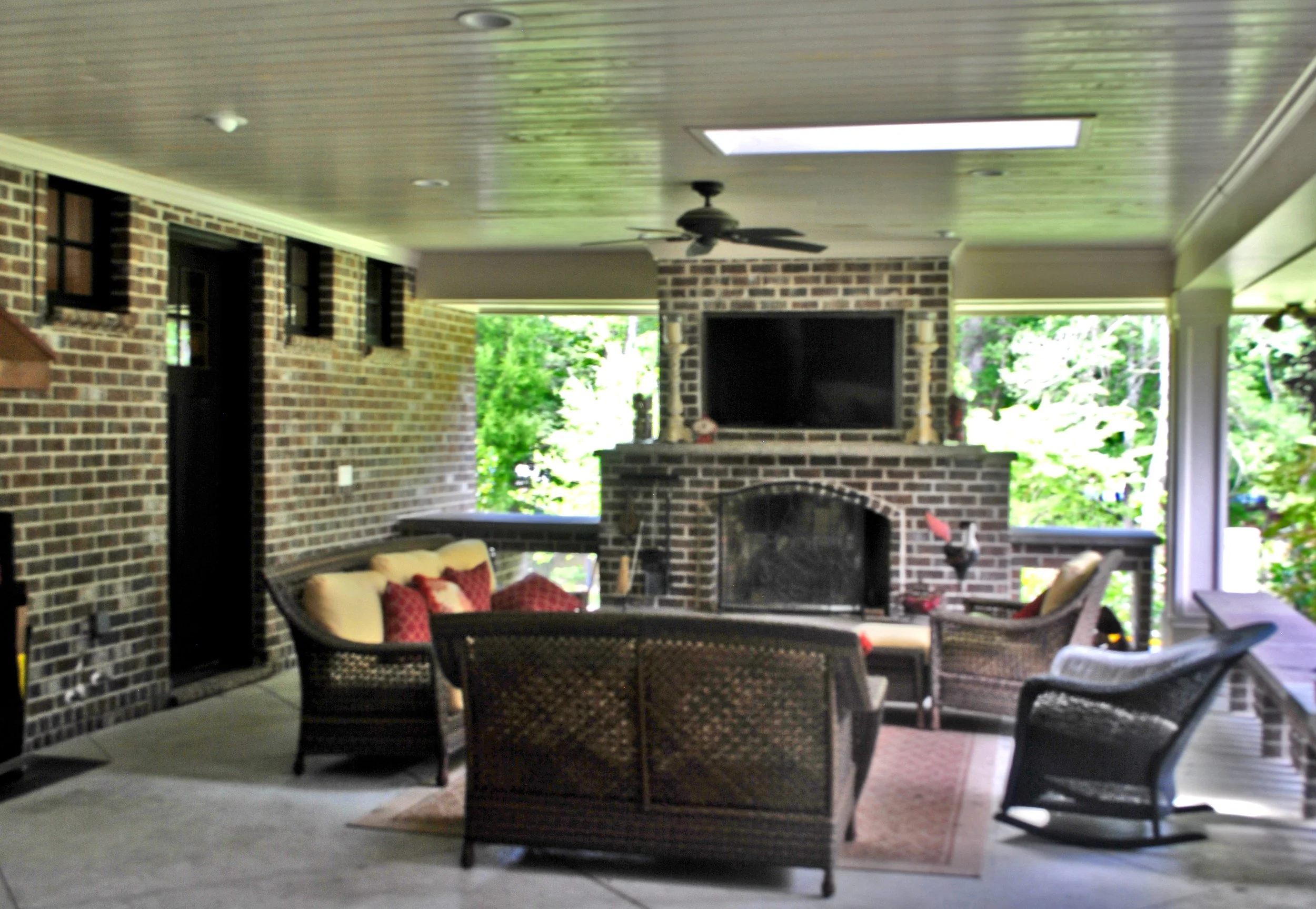 Covered outdoor porch area with wicker furniture and a brick fireplace, surrounded by large windows with views of green trees.