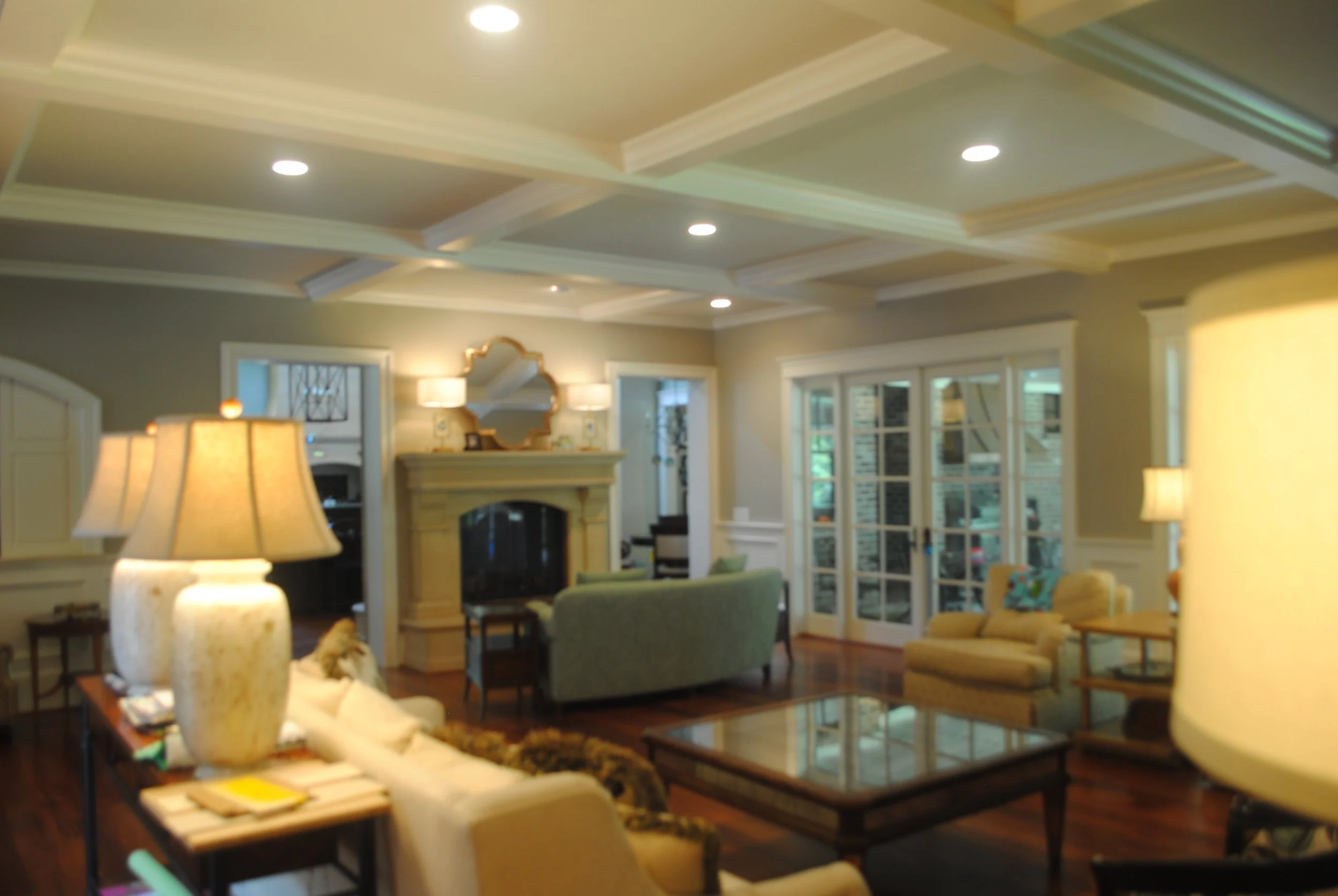 A cozy living room with a beige sofa, green armchair, glass coffee table, and tabletop lamps. There are windows with white framing and a fireplace with a mirror above it. Recessed ceiling lights illuminate the room.