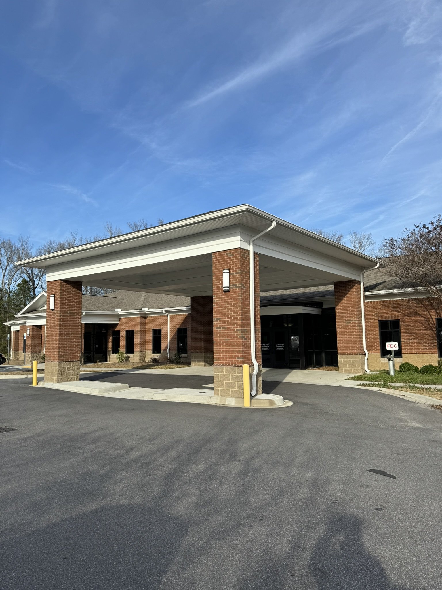 Carport with brick pillars and a gabled roof, parking area with yellow bollards, building with brick walls and large windows, trees in the background, and a bright blue sky with wispy clouds.