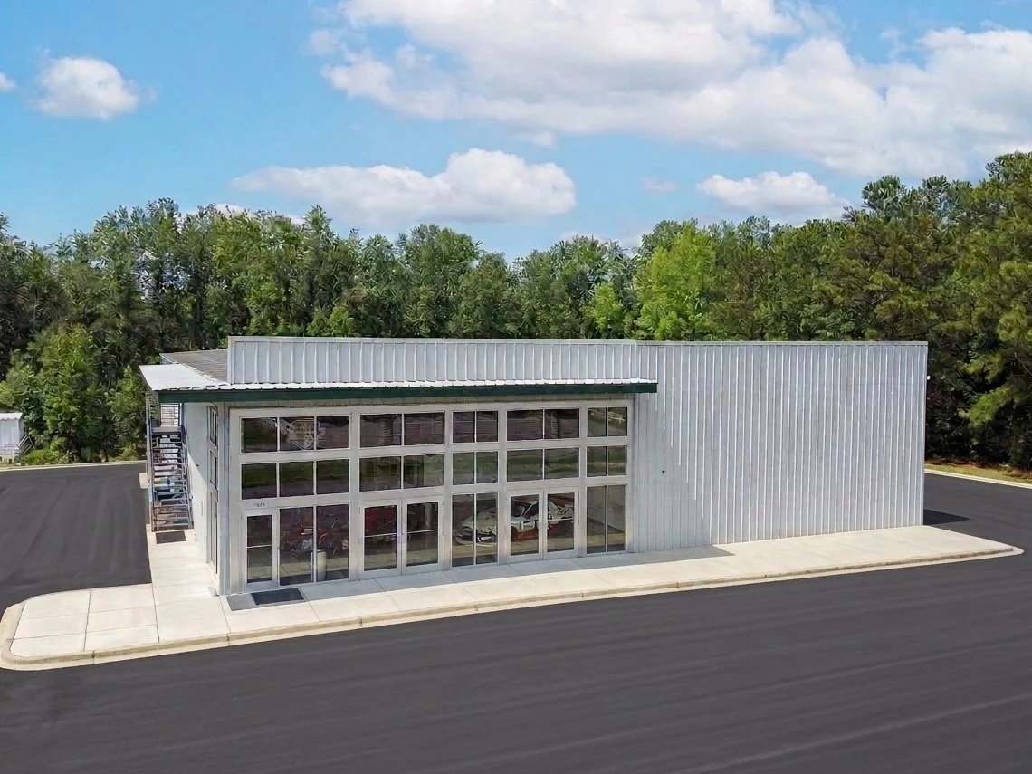 A modern commercial building with large glass windows and a metal exterior, situated on a paved lot with a sidewalk, surrounded by trees under a partly cloudy sky.