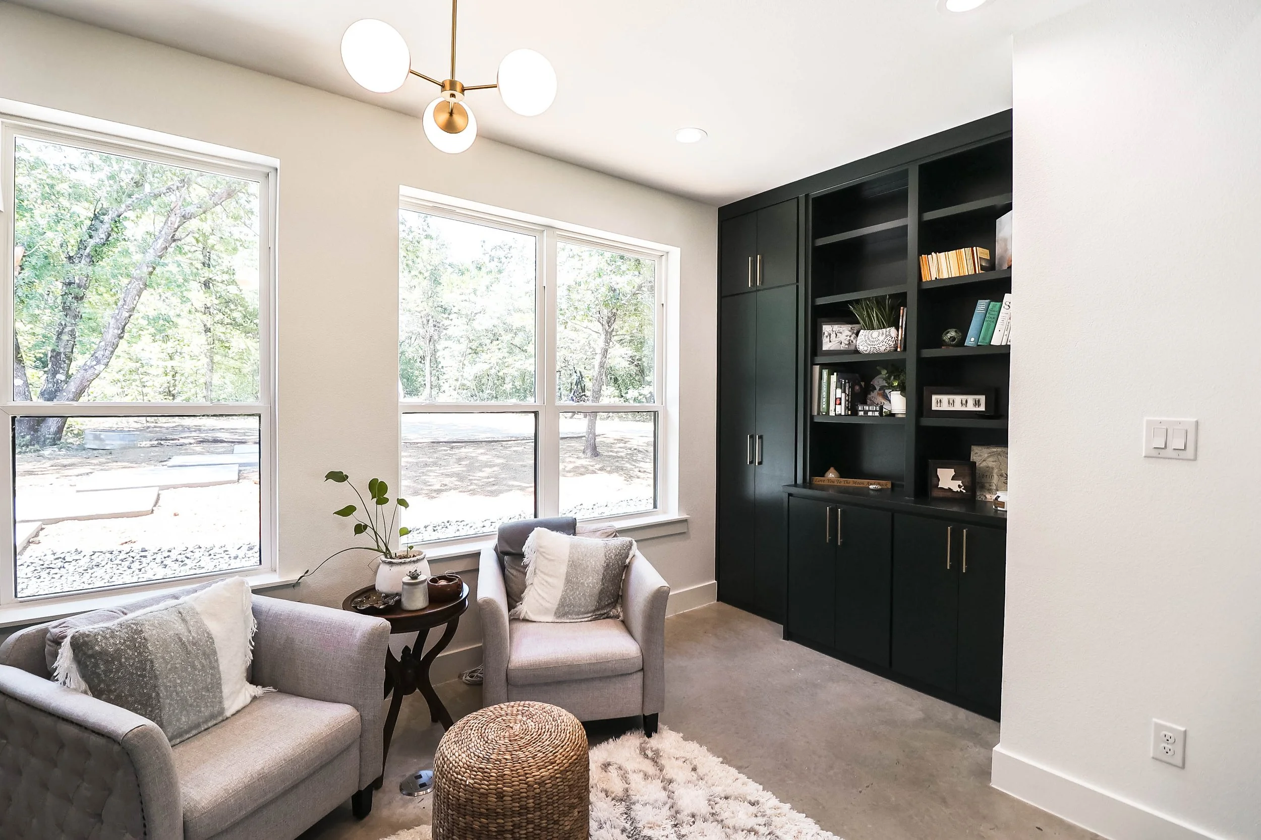 Living room with large windows, black built-in bookshelf, and cozy seating area with beige armchairs, throw pillows, a round woven ottoman, and a small side table with decorative items.