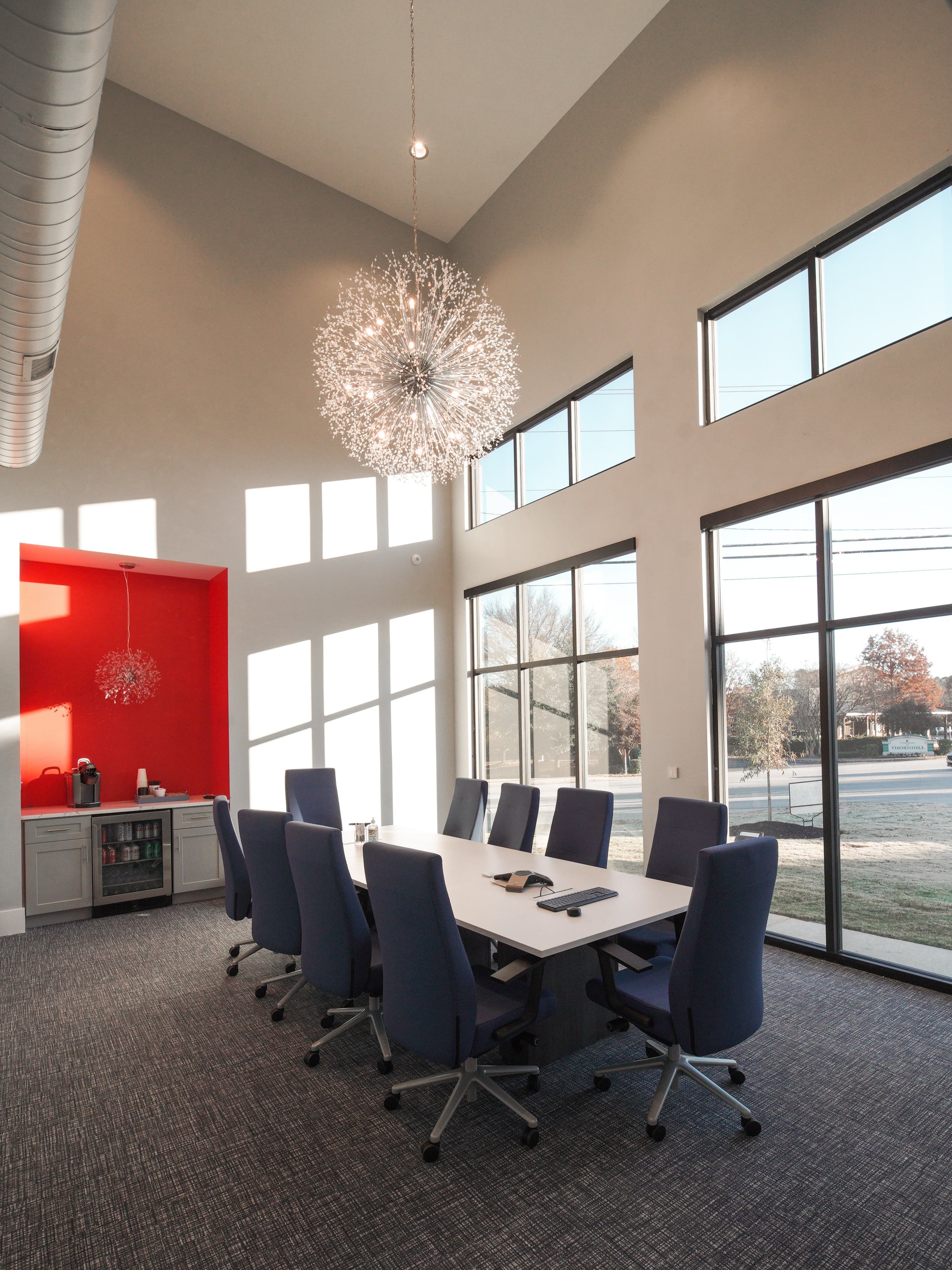 Modern conference room with large windows, a long white table, blue ergonomic chairs, dark carpet, a red accent wall with a small kitchen area, and a large spherical chandelier hanging from the ceiling.
