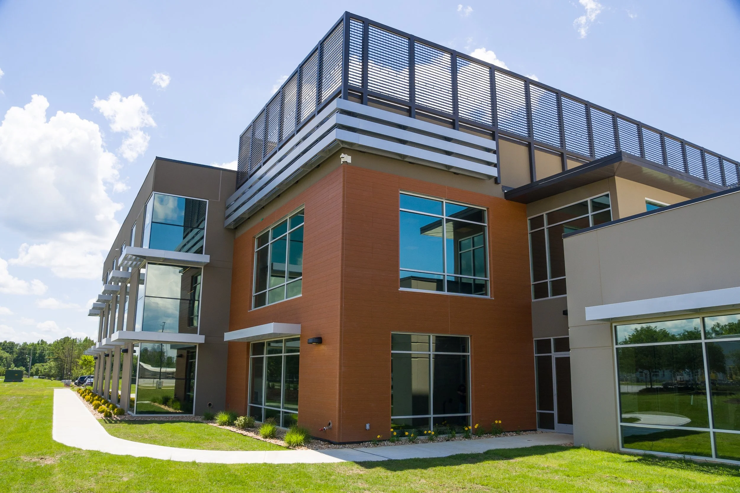 Modern multi-story residential or commercial building with large windows, orange and beige exterior walls, and a rooftop terrace with metal railing, set against a partly cloudy blue sky.