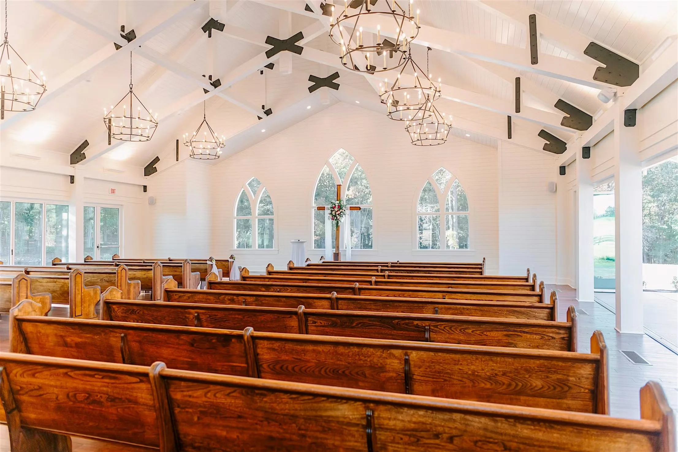 Interior of a church with wooden pews, large arched windows, and a cross with flower arrangement at the front. The ceiling has white wood paneling, black air vents, and hanging chandeliers with lit candles.