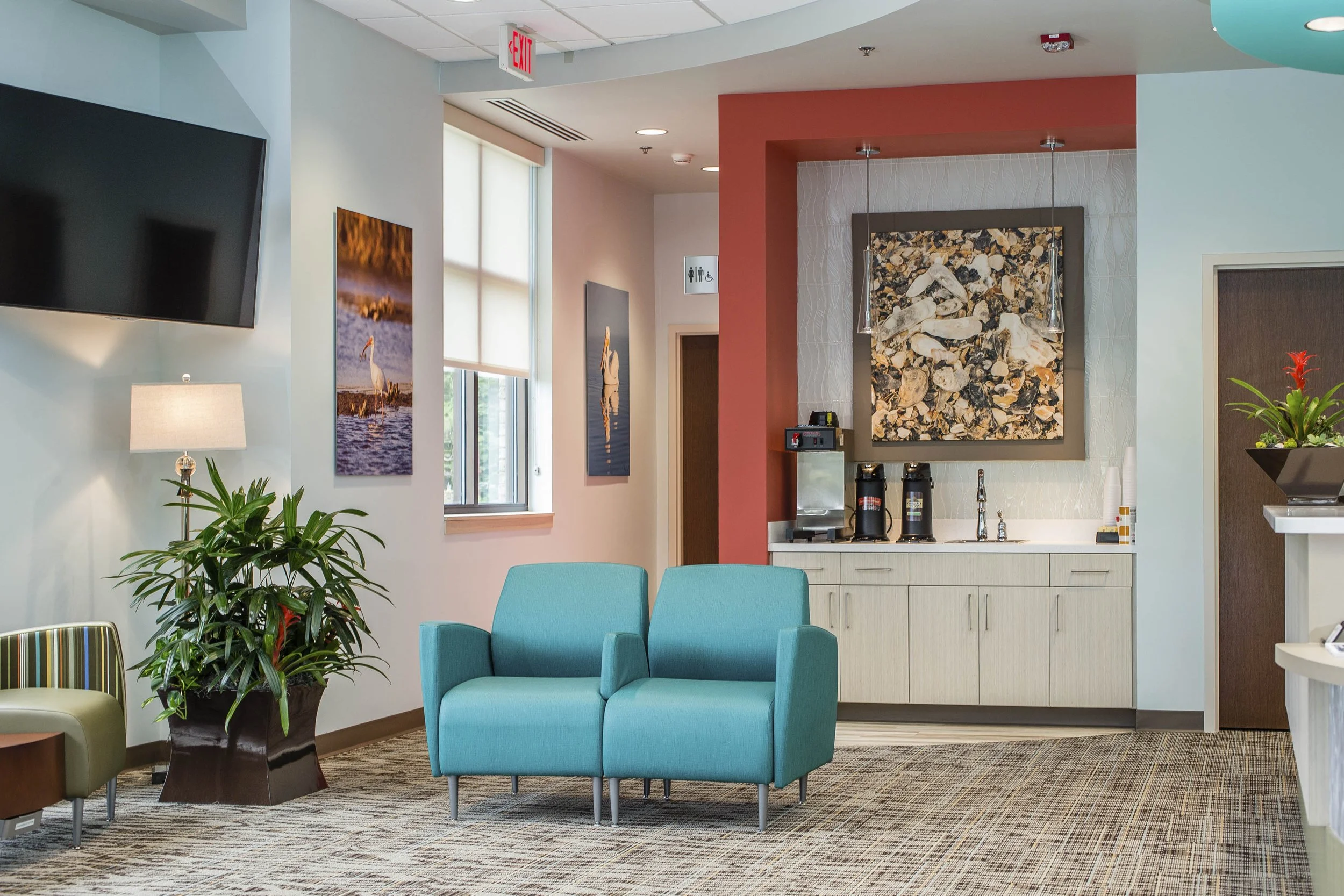 Interior of a modern waiting or lounge area with blue armchair, potted plant, wall-mounted television, framed art, and a kitchenette with a sink and coffee dispensers. Natural light coming through windows, and colorful wall art present.