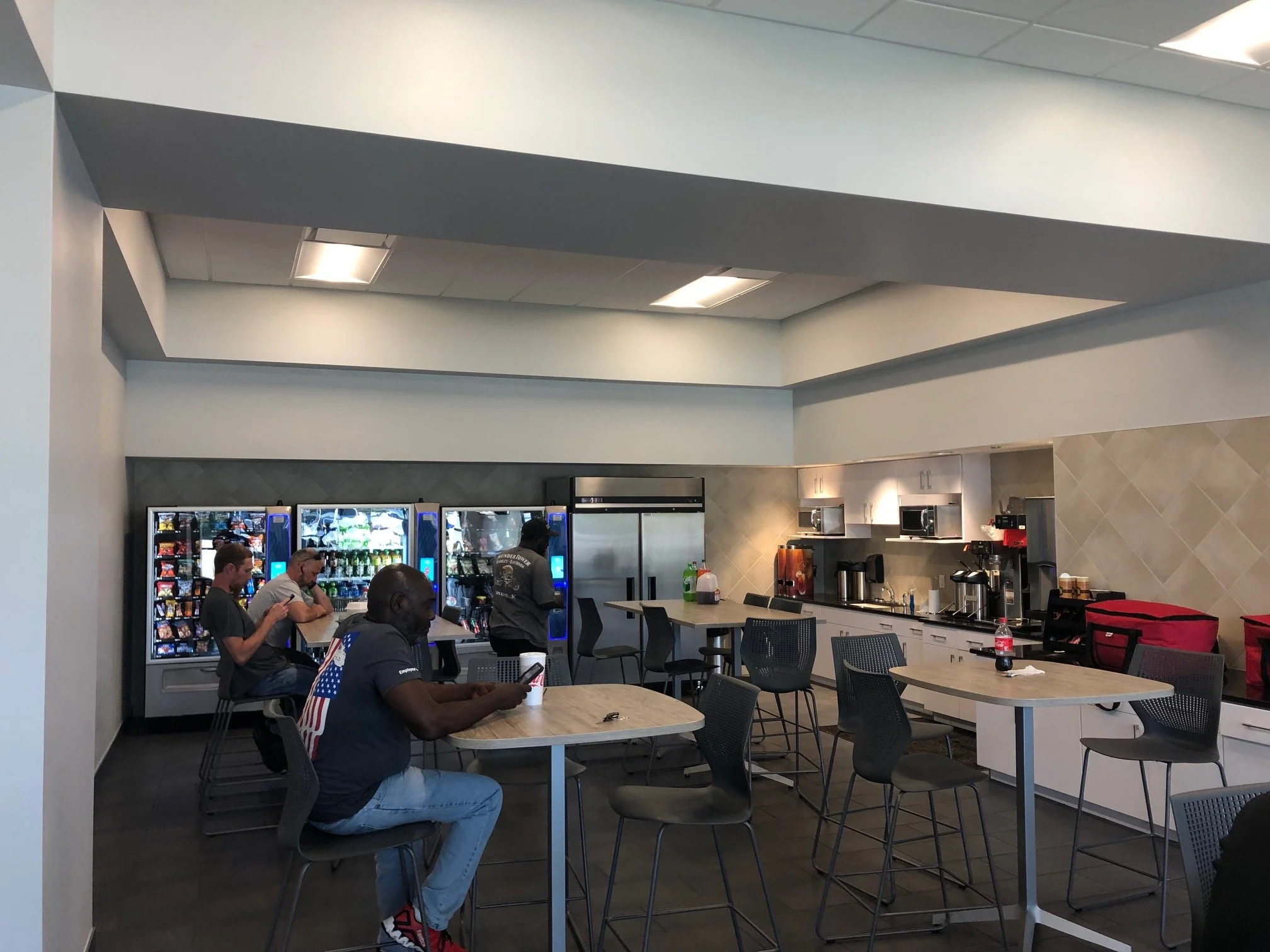 Inside a casual dining area with several people sitting at tables, some looking at their phones. In the background, there's a row of vending machines and a small kitchen area with appliances.