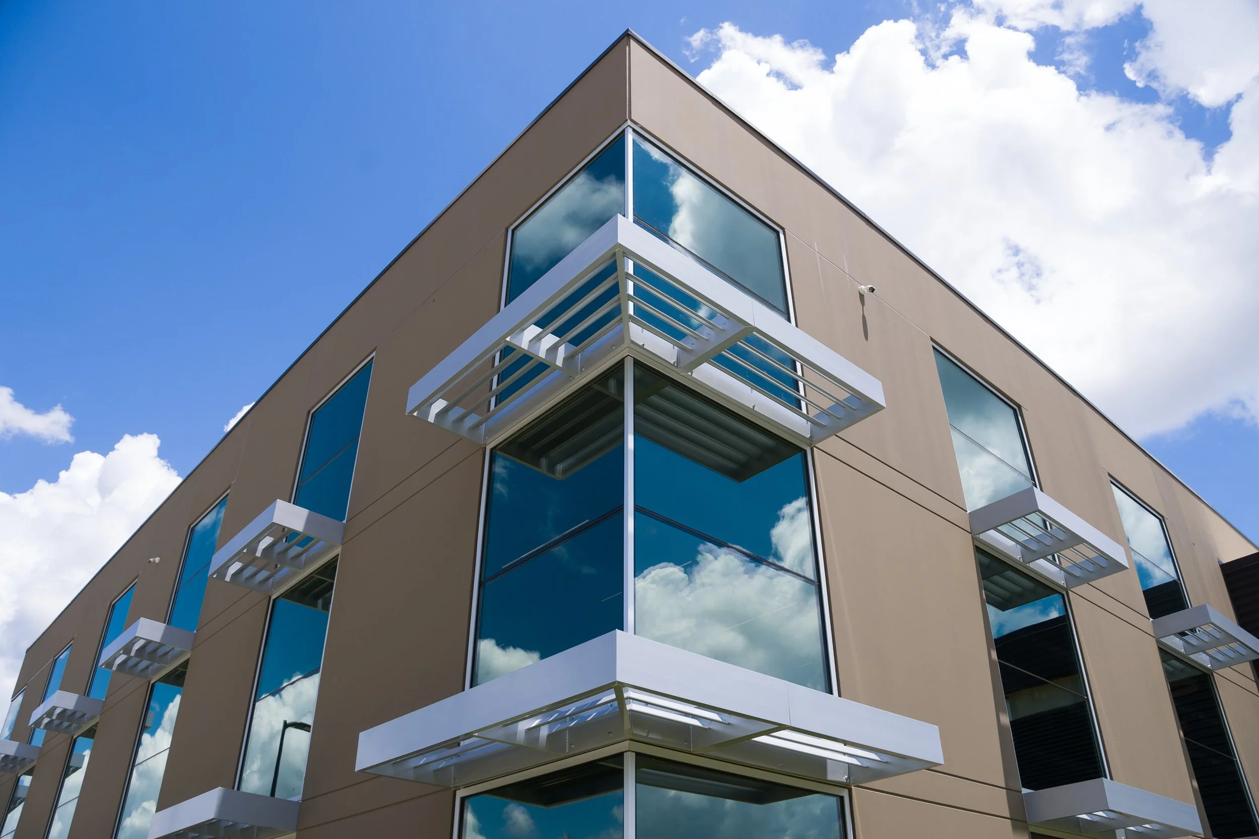 Modern beige building with large reflective glass windows and white balconies, under a partly cloudy blue sky.
