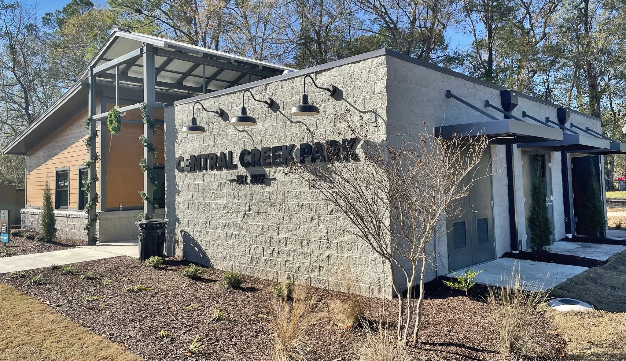 A modern building with a sign reading 'Central Creek Park' and the establishment year 2022. The building has exterior lights, a small landscaped garden with bushes and a leafless tree, and sits under a partly cloudy blue sky with trees in the background.