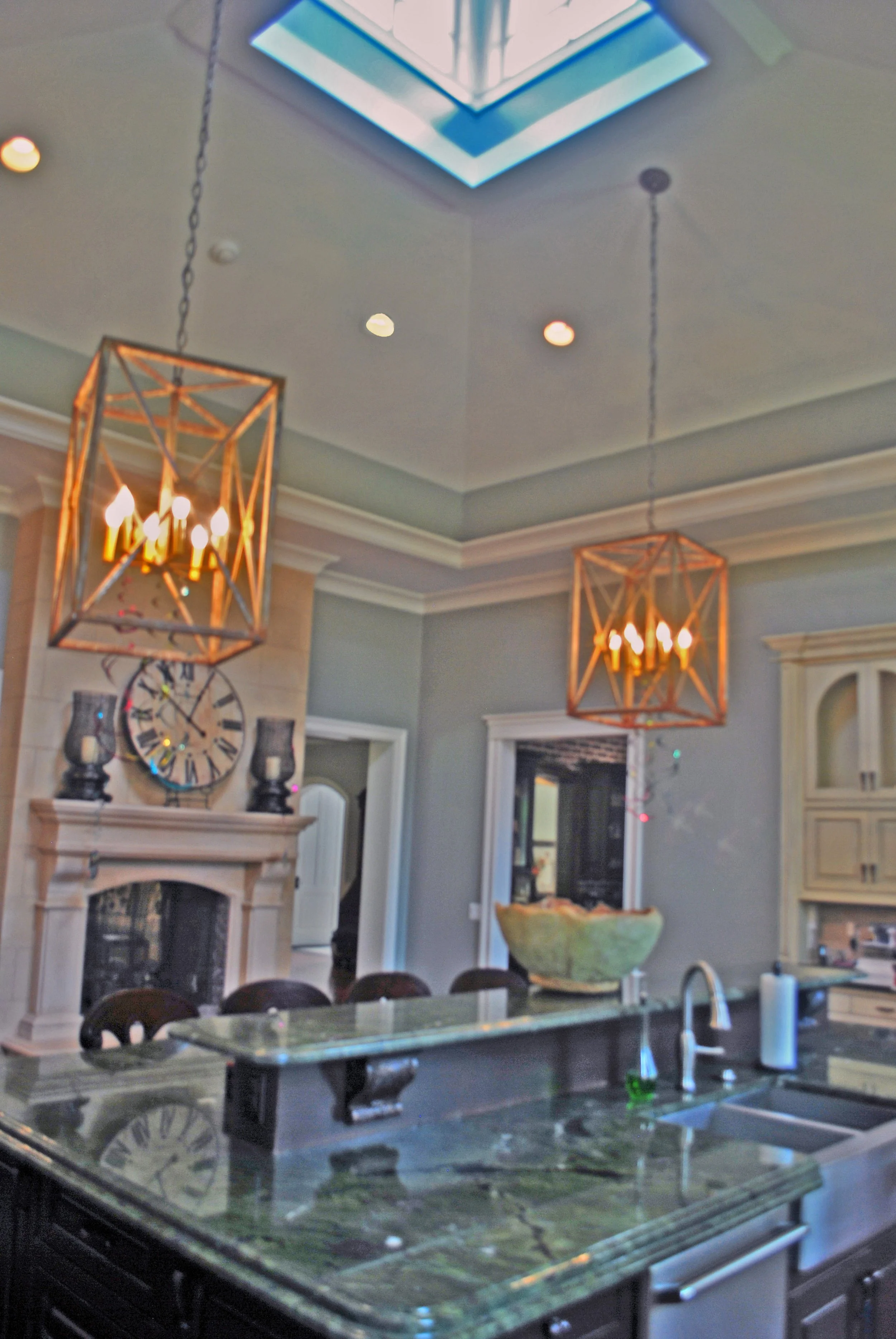 Kitchen with a green marble countertop, a large clock on the wall, and two hanging geometric chandeliers with candle-like lights. The ceiling has a skylight, and there are various decorative items like vases and a fireplace mantle in the background.