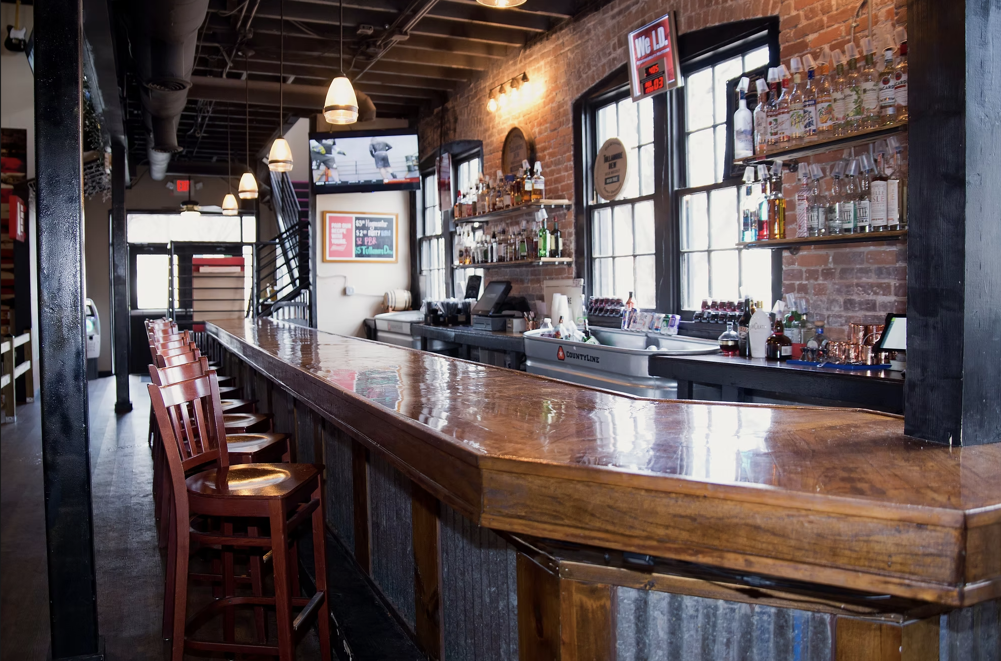 Interior of a bar or restaurant with a long polished wooden counter, red chairs, brick walls, large windows, hanging pendant lights, and shelves with liquor bottles.