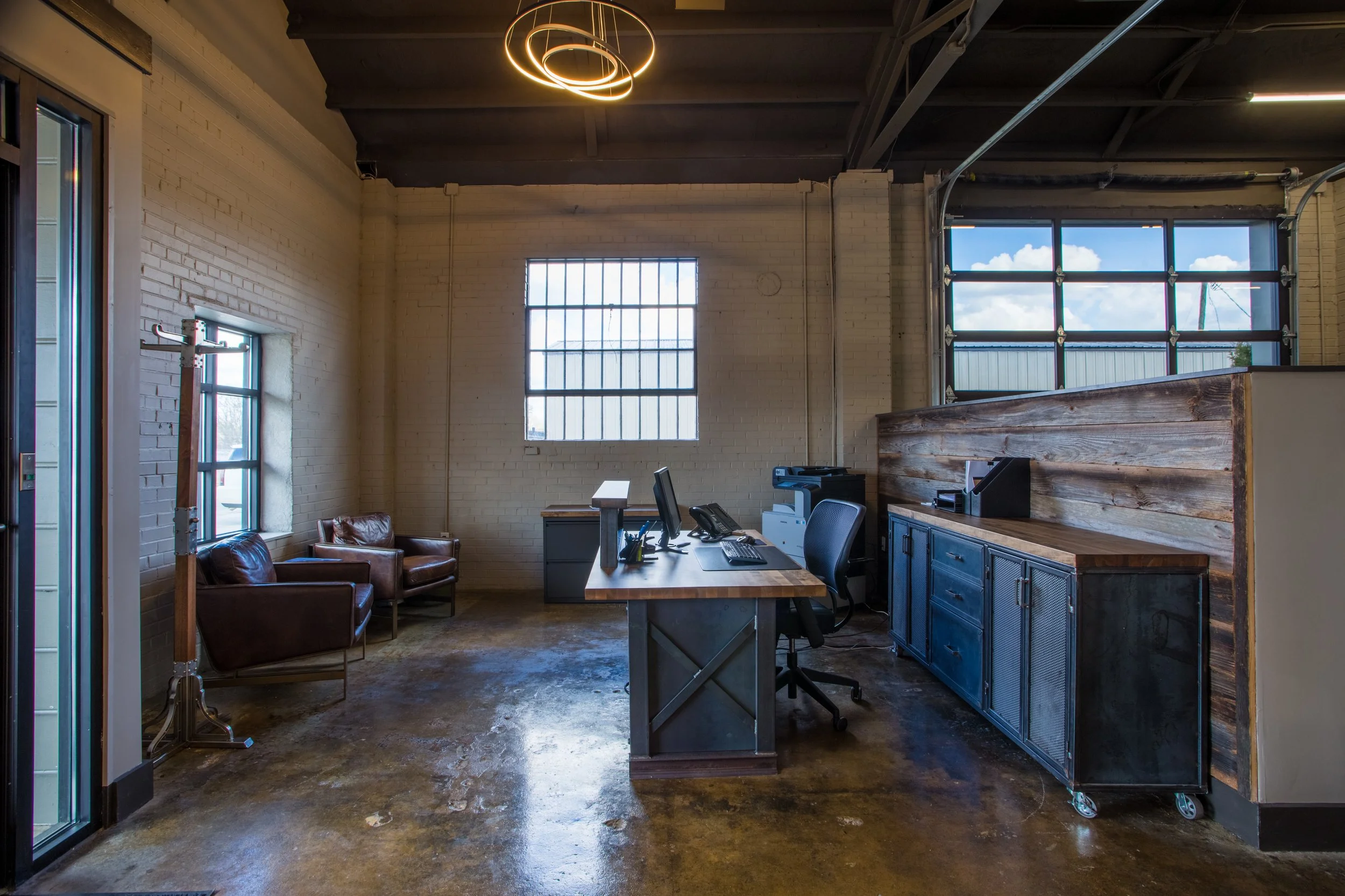 Industrial-style office space with high ceilings, large windows, a desk with office equipment, a leather sofa, and modern circular ceiling lights.