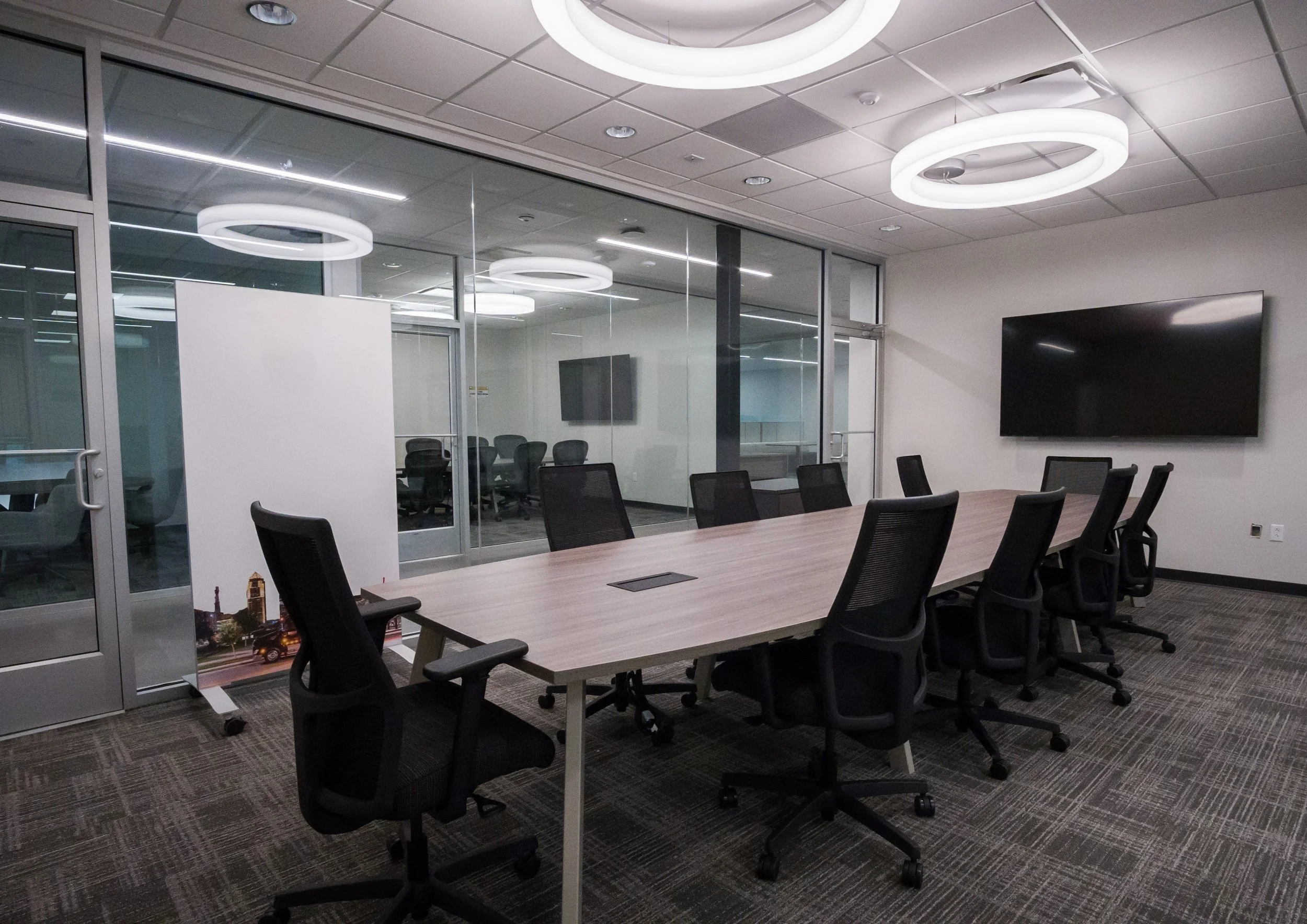 A modern conference room with a long wooden table, black ergonomic chairs, glass walls, and circular LED ceiling lights, with a large flat-screen monitor on the wall.