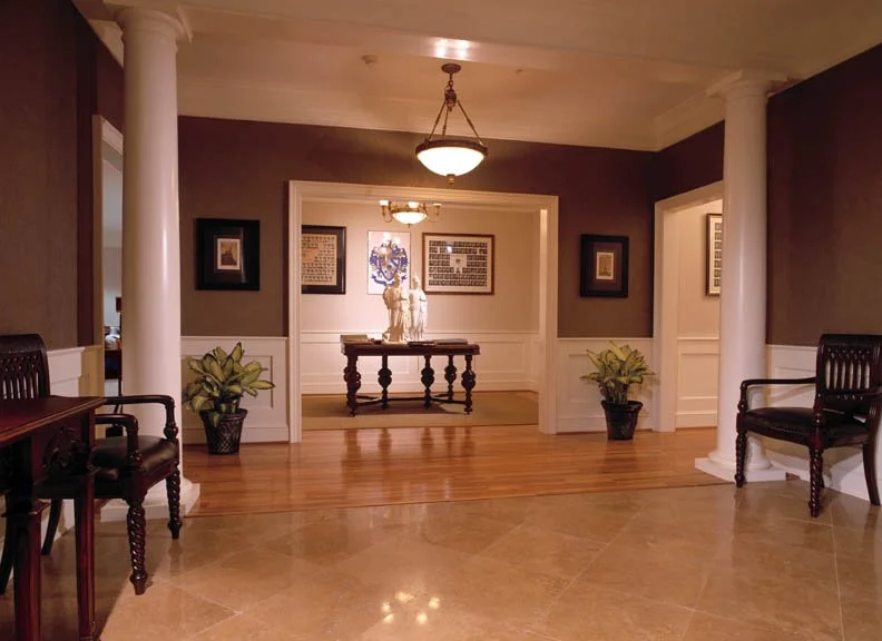 Entrance hall of a home with wooden flooring, two black leather chairs, two potted plants, and a visible doorway leading to a dining room with a table, a sculpture on it, framed pictures, and chandeliers.